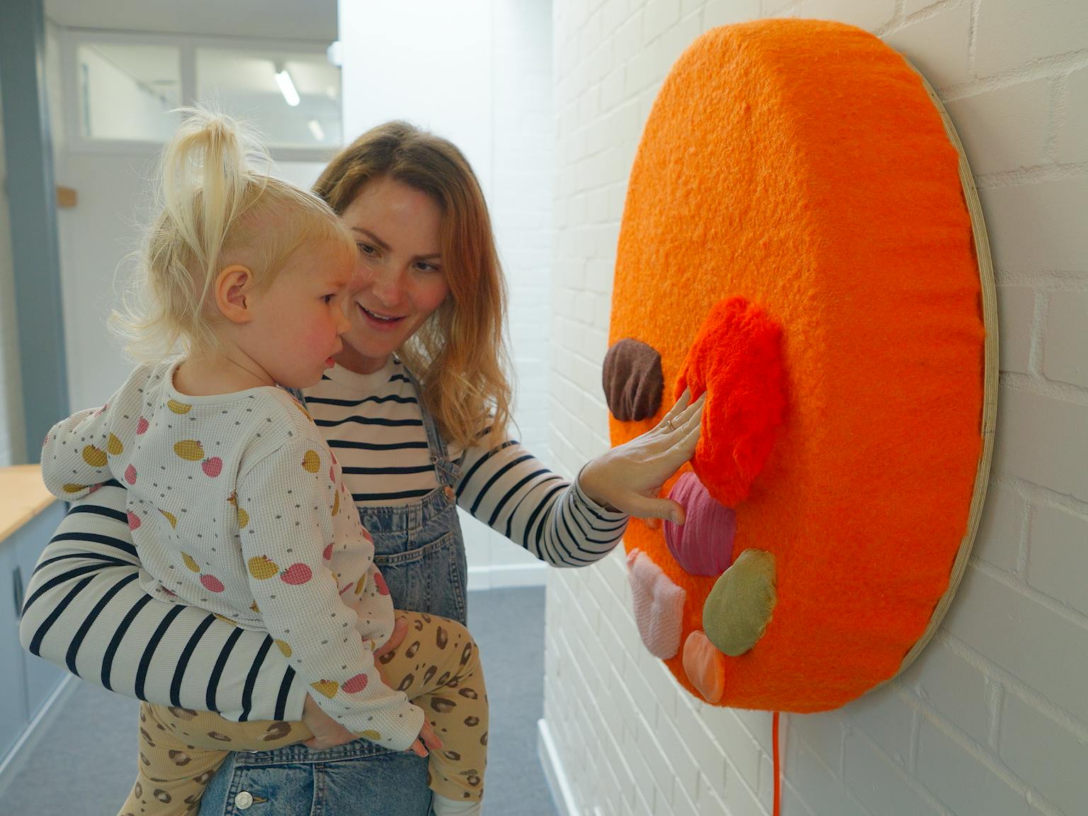 A mother and infant engaging with an installation. The mother is touching an orange circle with different colour shapes and textures on it.