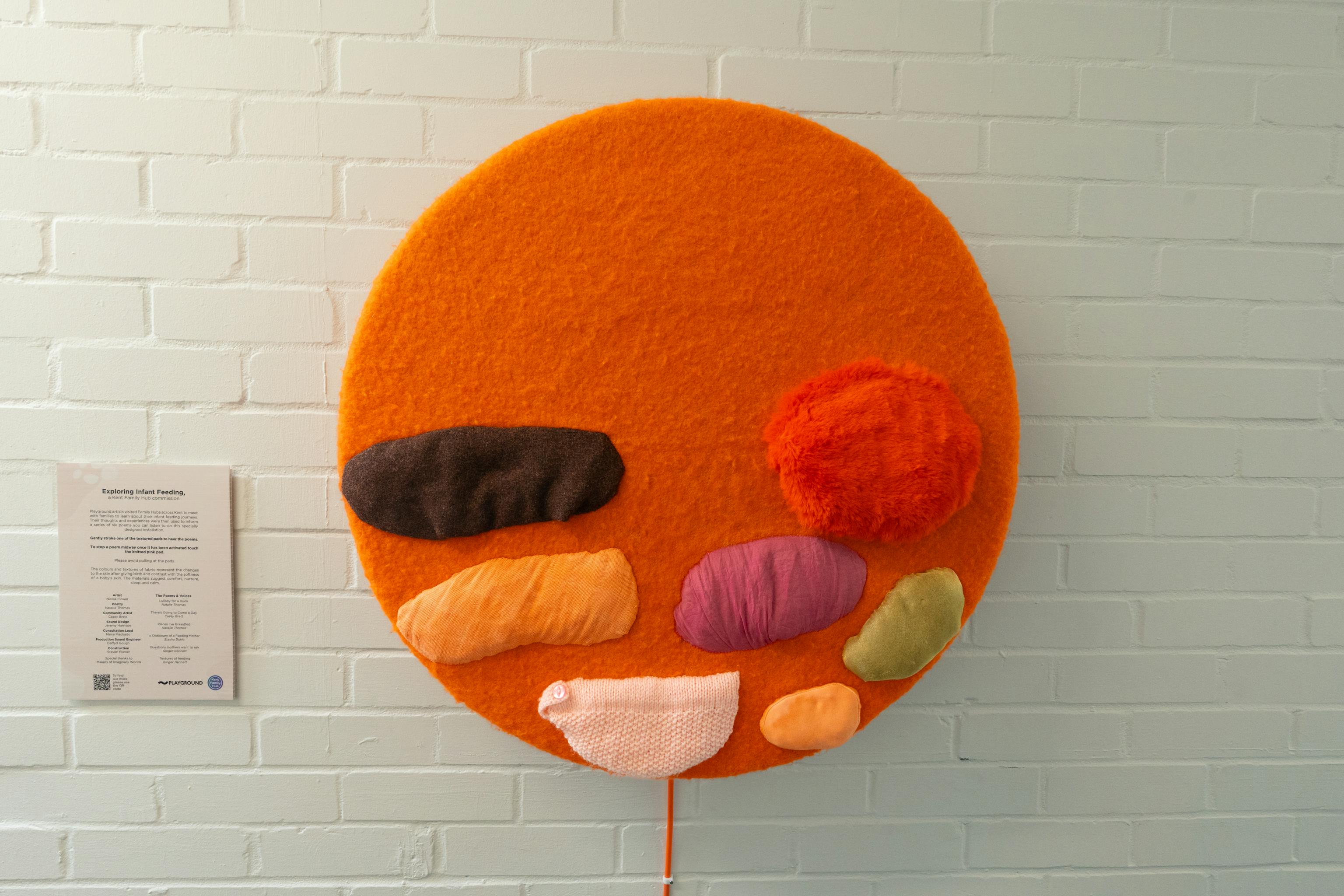 An art installation of the Infant Feeding project on display at a Family Hub. The artwork is a textured orange circle with different coloured shapes and textures added to it. It is displayed against a white wall. Next to it is some text explaining the artwork