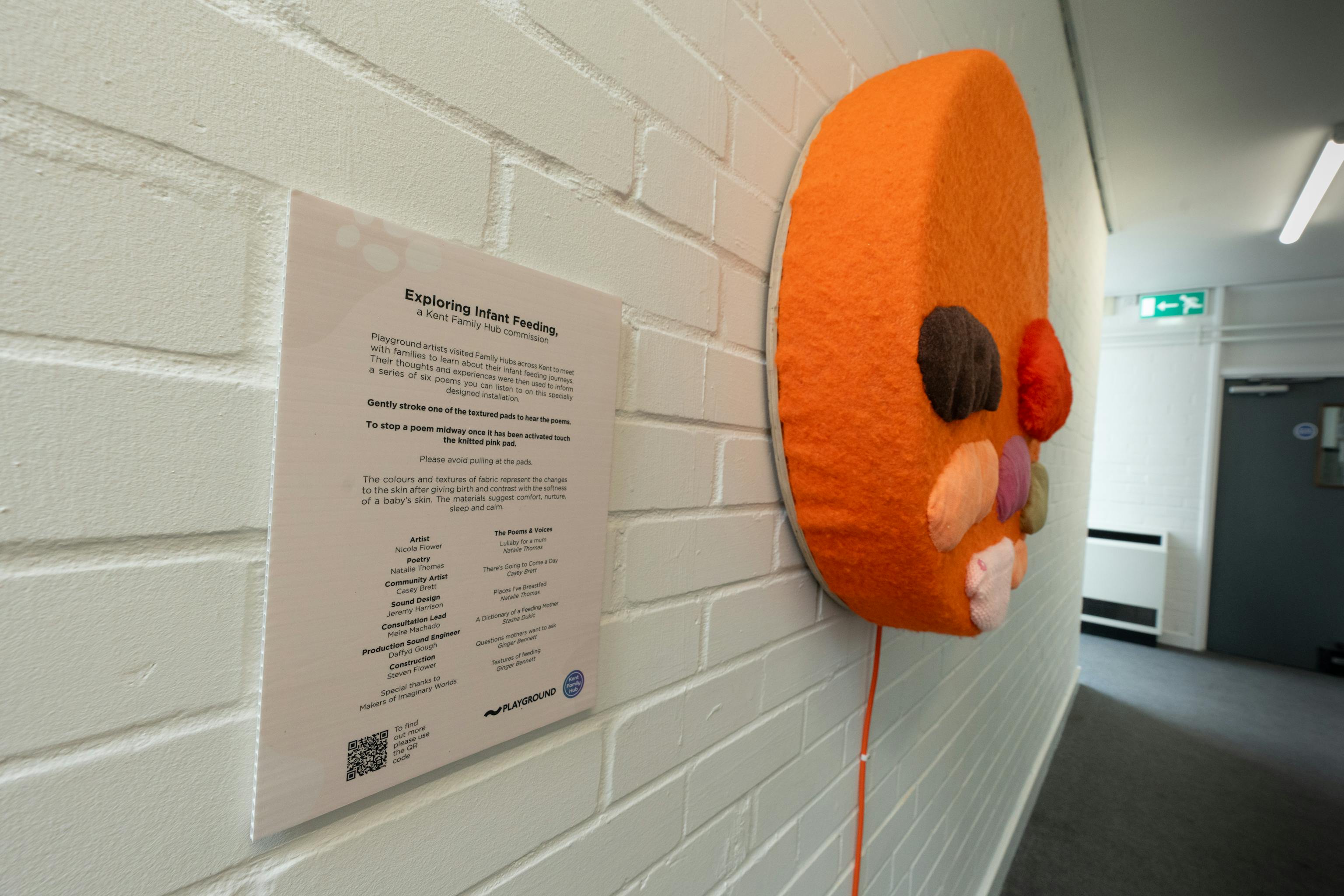 An art installation of the Infant Feeding project on display at a Family Hub. The artwork is a textured orange circle with different coloured shapes and textures added to it. It is displayed against a white wall. Next to it is some text explaining the artwork