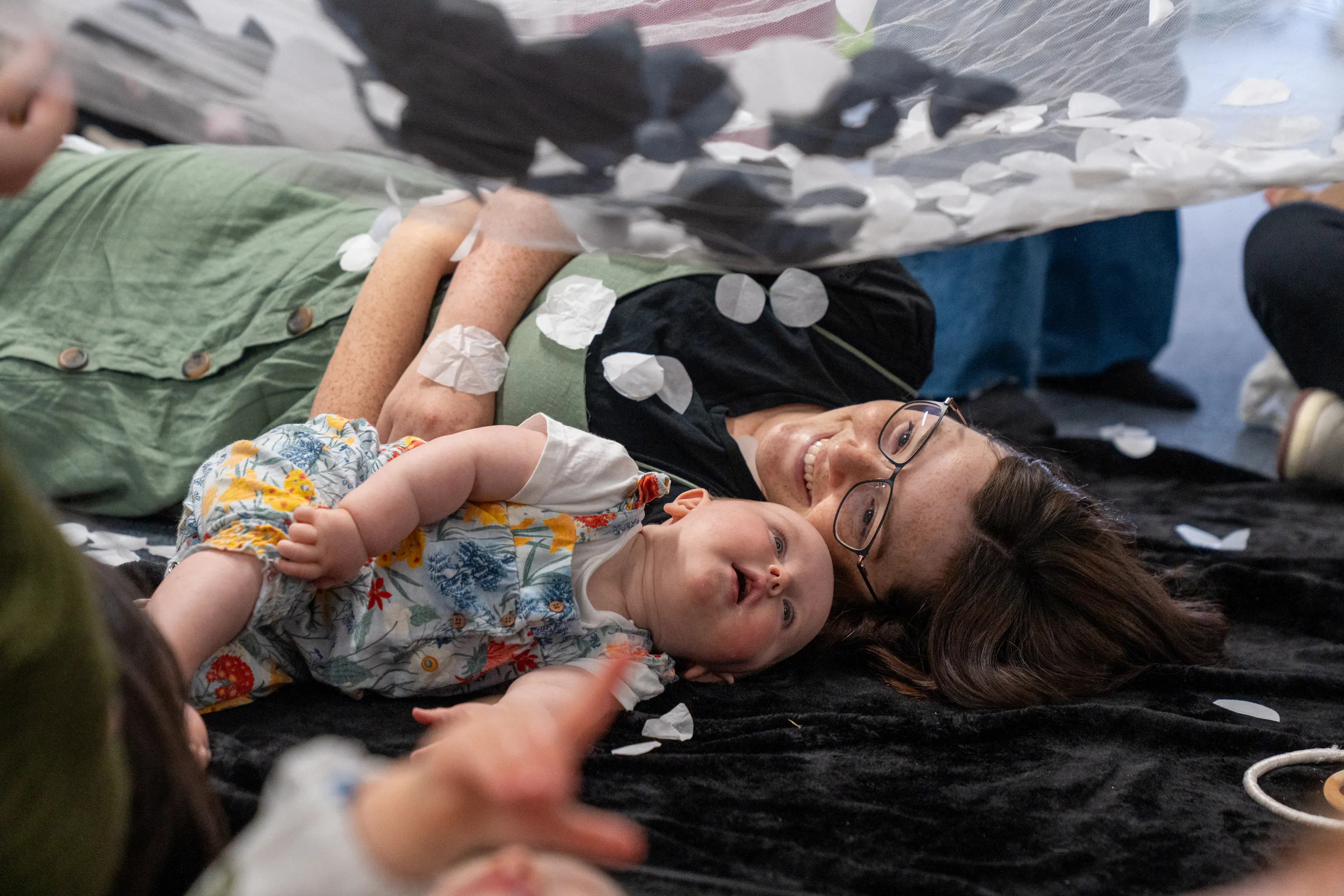 A baby and mother are lying next to each other on a textured black mat. The mother is smiling at her baby and the baby is looking up at a net with black and white materials on it