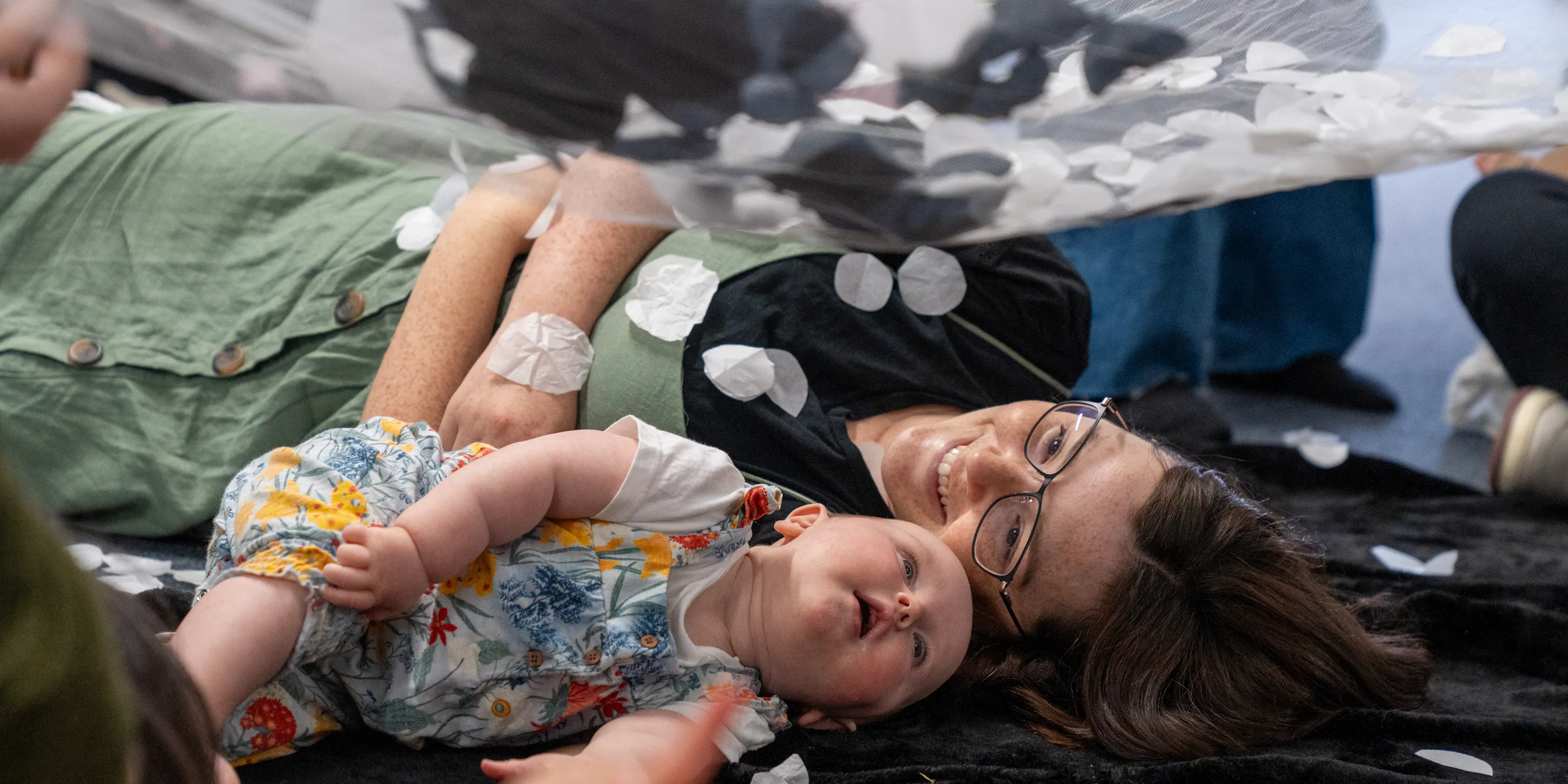 A baby and mother are lying next to each other on a textured black mat. The mother is smiling at her baby and the baby is looking up at a net with black and white materials on it