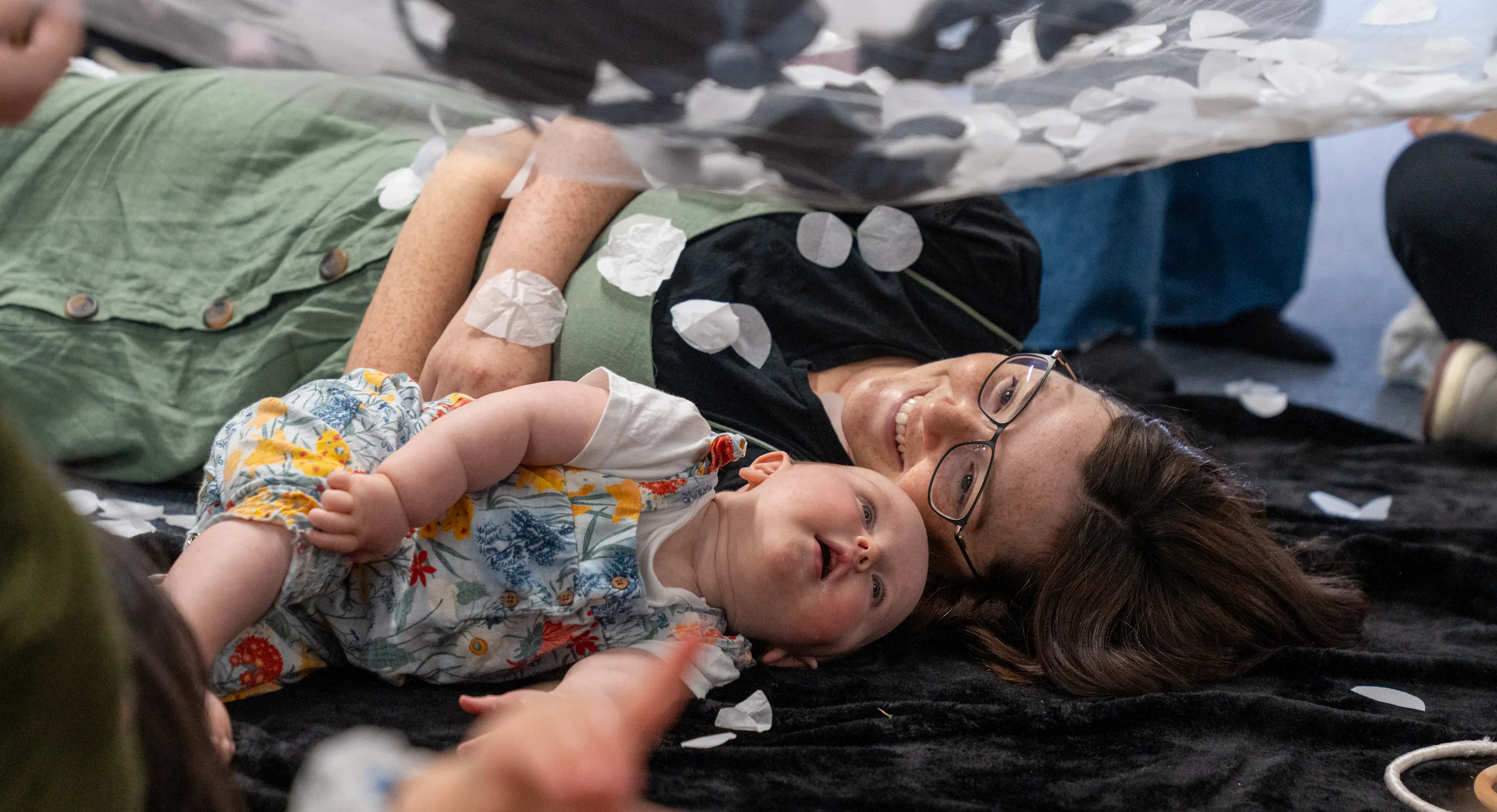A baby and mother are lying next to each other on a textured black mat. The mother is smiling at her baby and the baby is looking up at a net with black and white materials on it