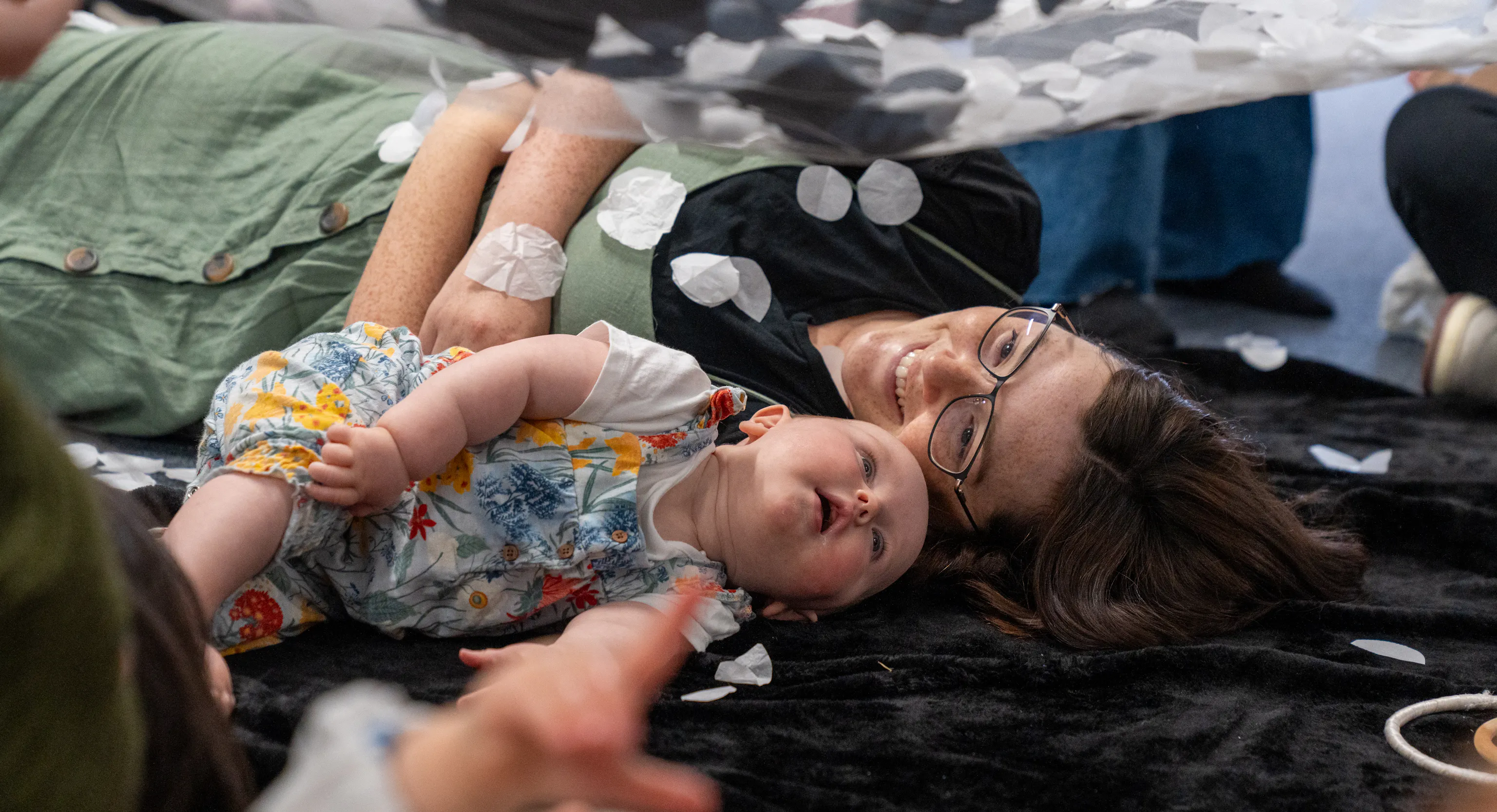 A baby and mother are lying next to each other on a textured black mat. The mother is smiling at her baby and the baby is looking up at a net with black and white materials on it