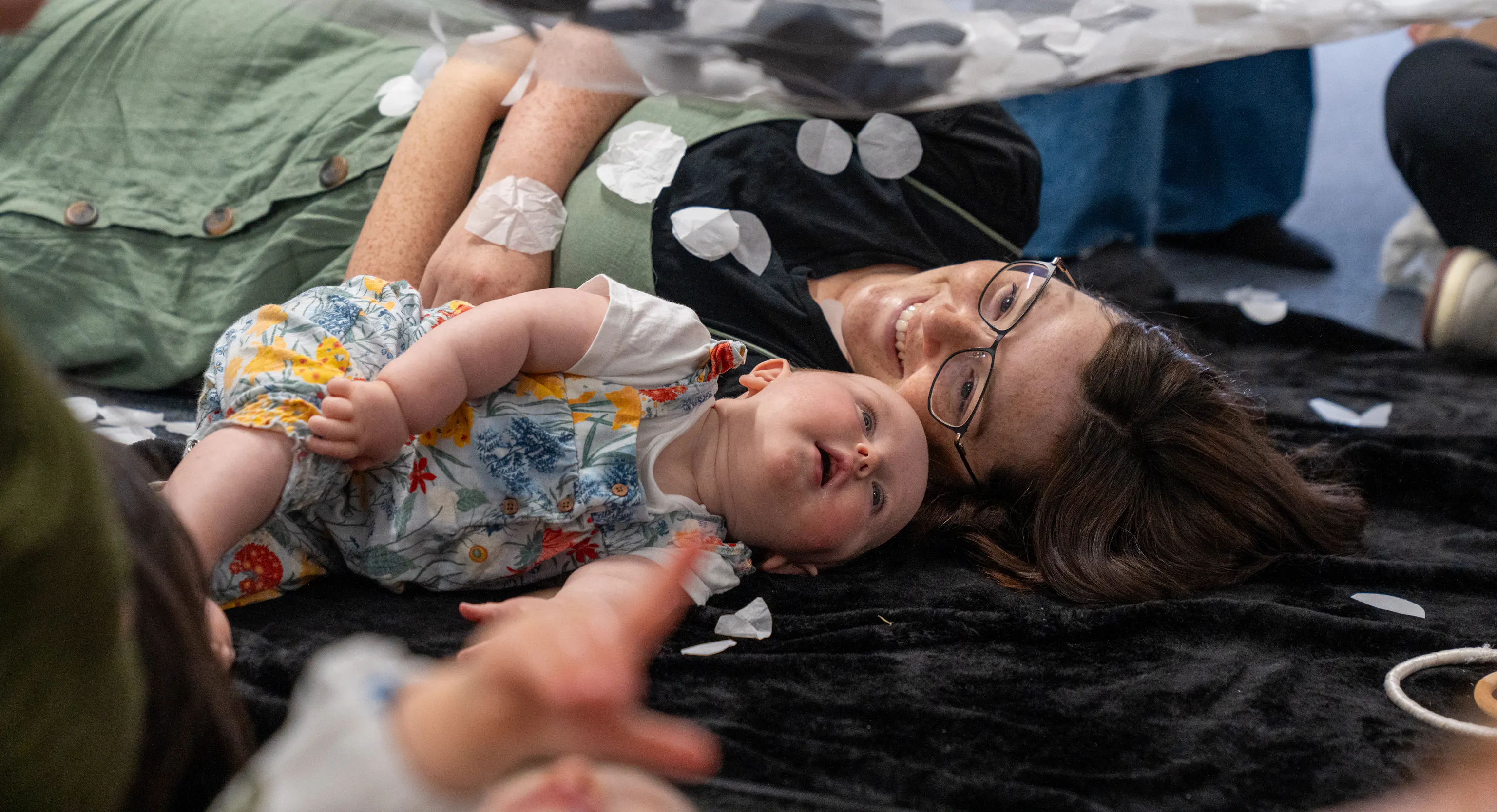 A baby and mother are lying next to each other on a textured black mat. The mother is smiling at her baby and the baby is looking up at a net with black and white materials on it
