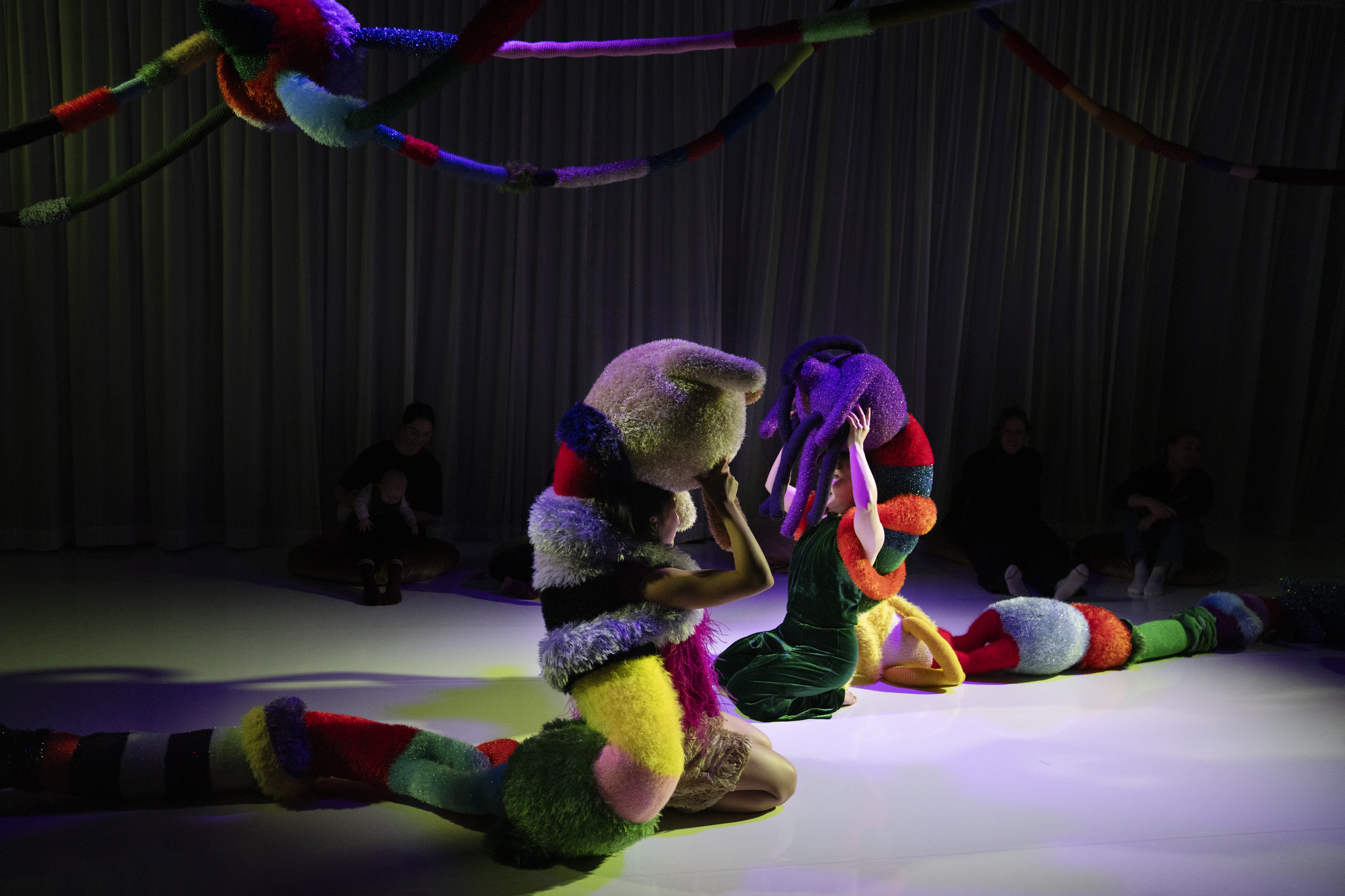 Two creatures made from colourful and fluffy textures sit in a darkly lit room 