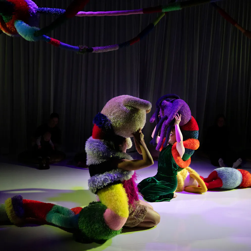 Two creatures made from colourful and fluffy textures sit in a darkly lit room