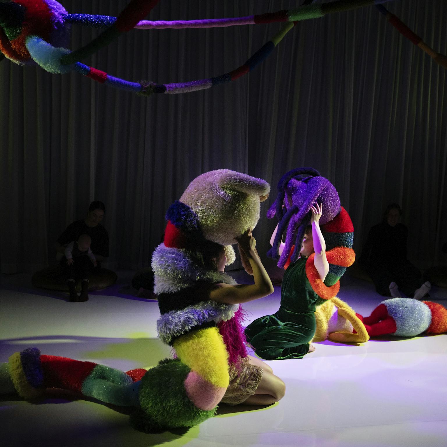 Two creatures made from colourful and fluffy textures sit in a darkly lit room