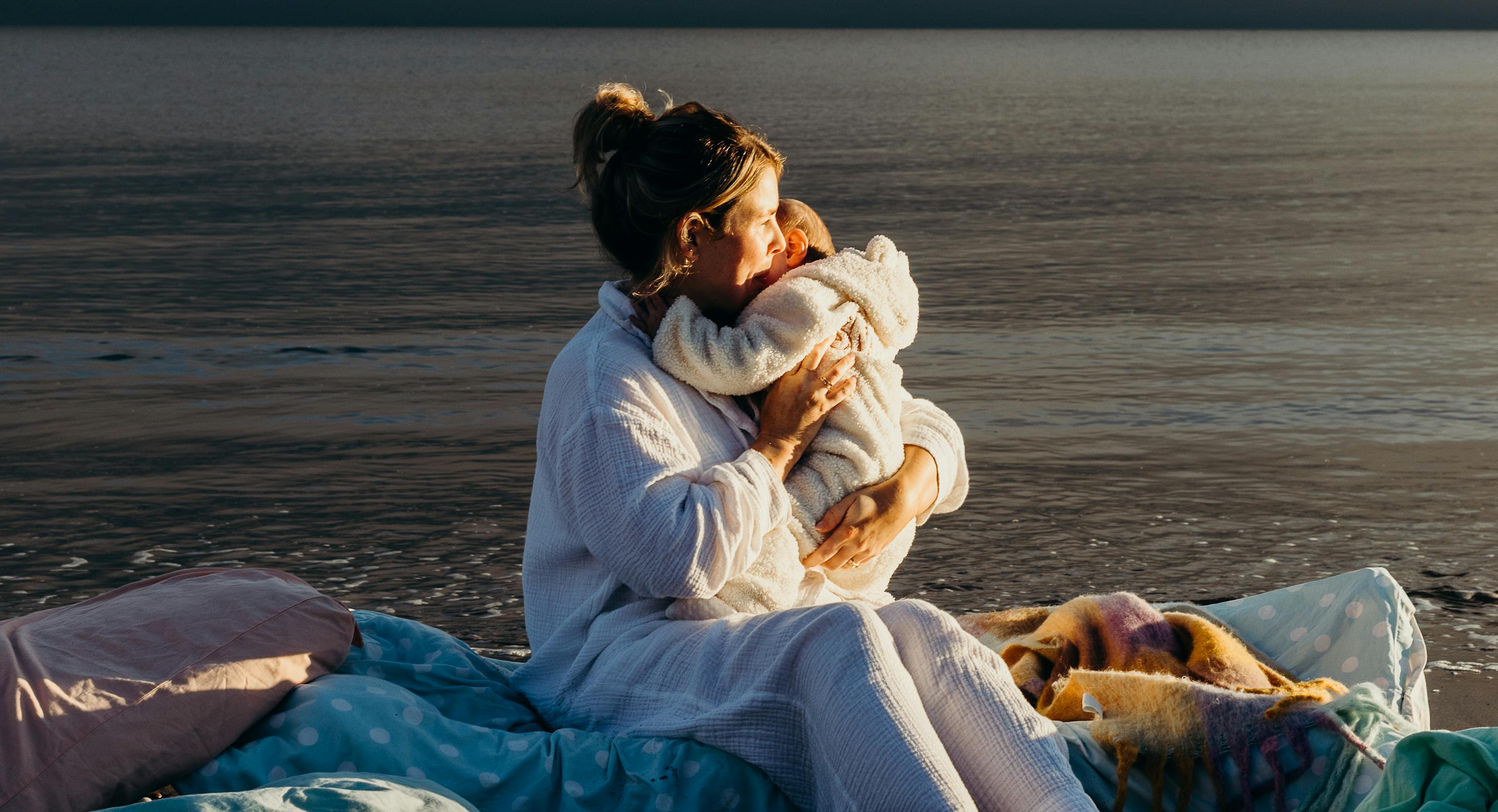A baby and mother embracing. In the background is the sea