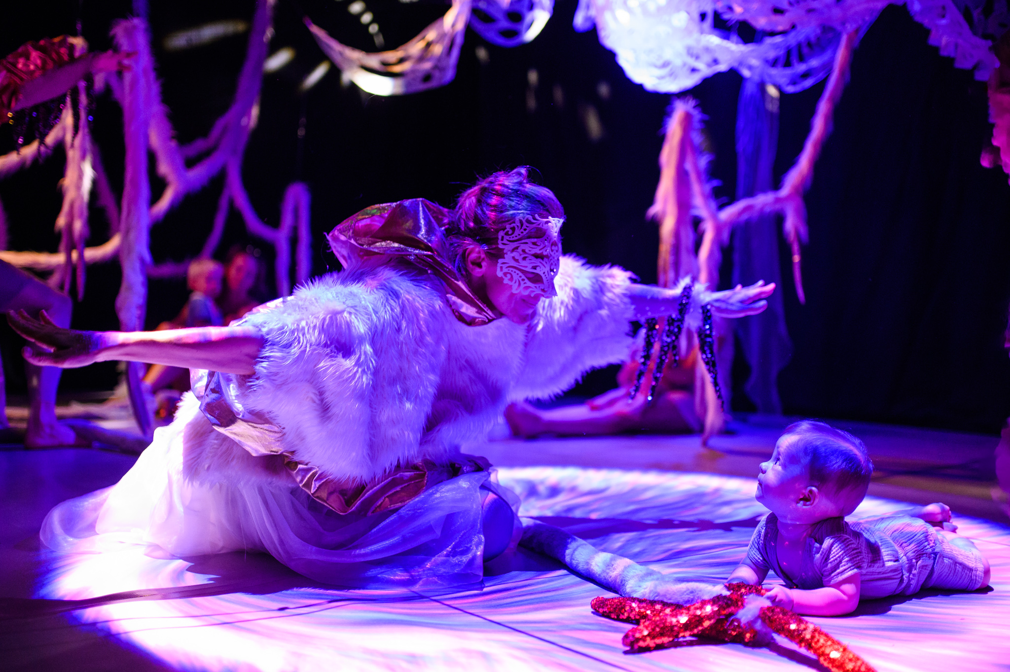 A performer wearing a fluffy feather top and a mask has their hands wide open in front of a baby lying on their front. The background is dark with textured materials hanging from the ceiling 