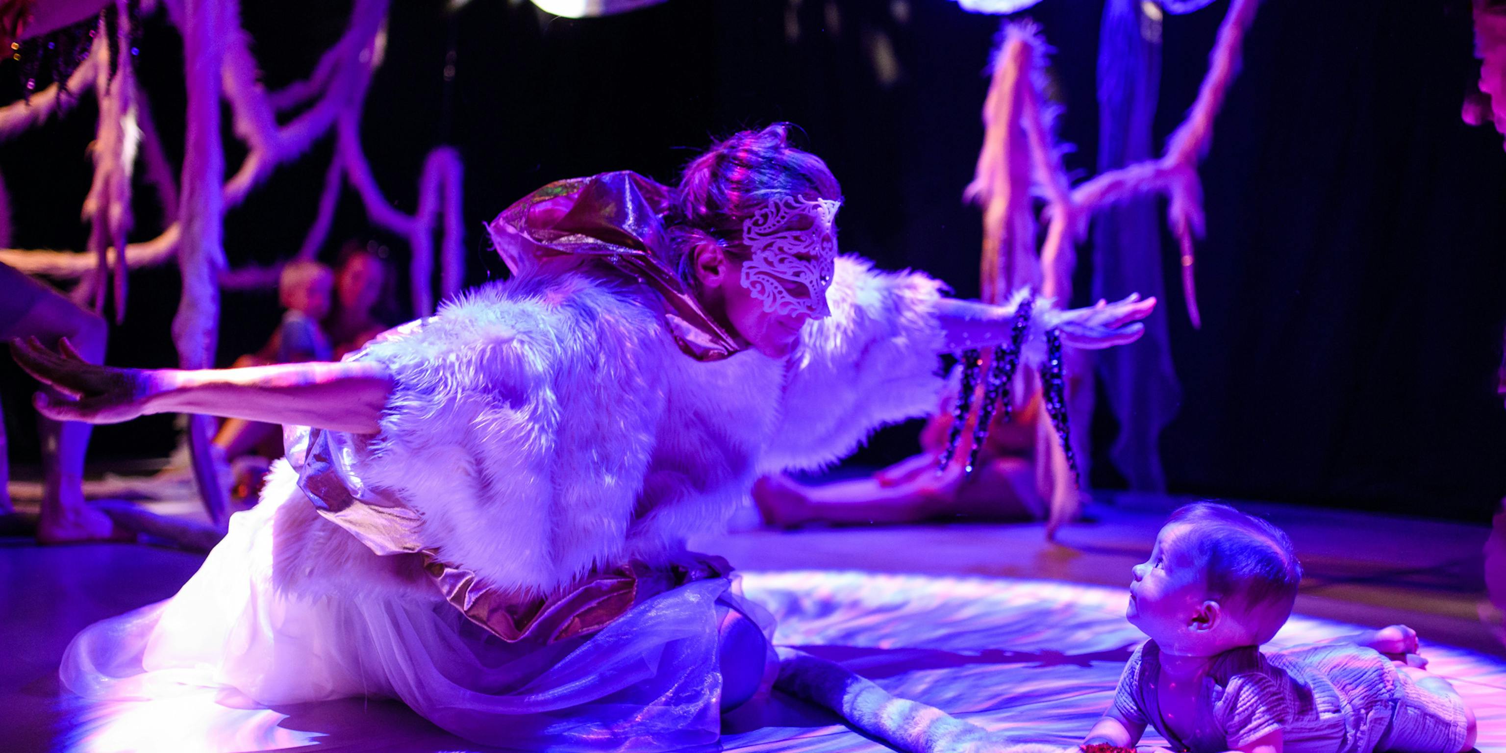 A performer wearing a fluffy feather top and a mask has their hands wide open in front of a baby lying on their front. The background is dark with textured materials hanging from the ceiling
