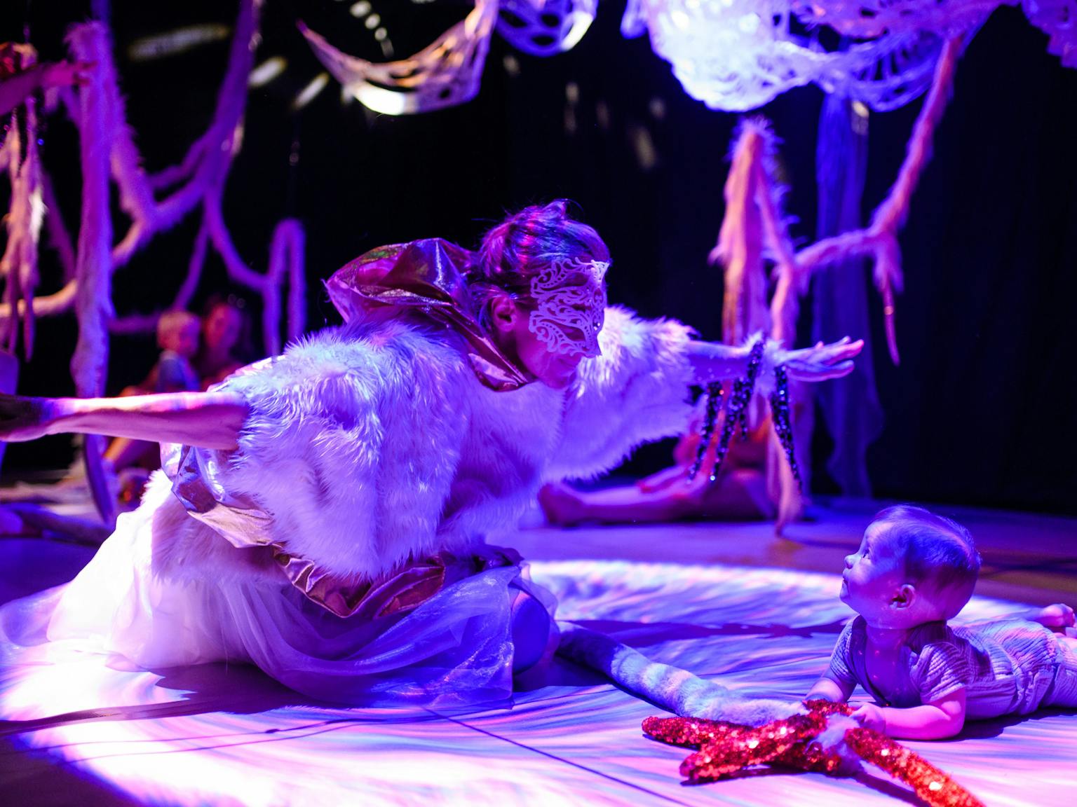 A performer wearing a fluffy feather top and a mask has their hands wide open in front of a baby lying on their front. The background is dark with textured materials hanging from the ceiling