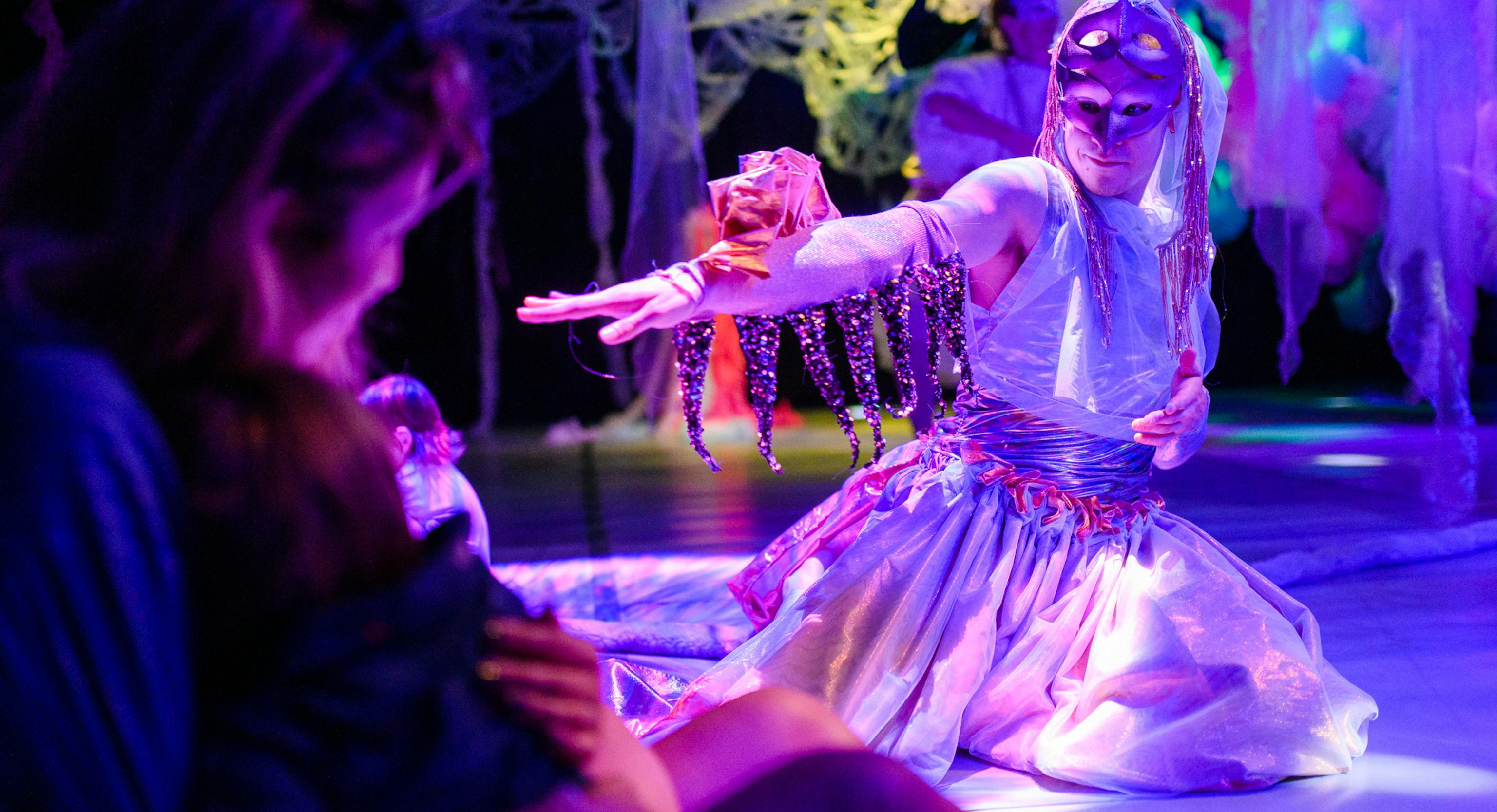 A parent and baby are watching a performer. The performer is wearing a mask and kneeling on the floor mid performance. They have material on the top of their hand and sequins hanging down.