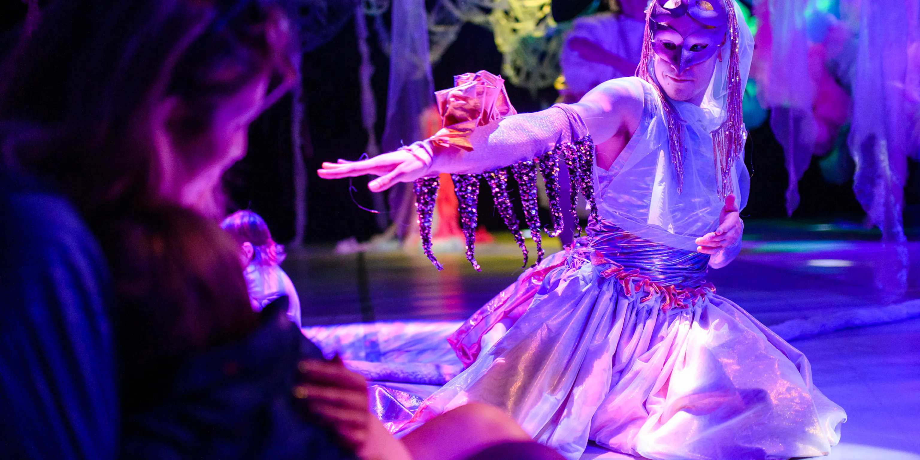 A parent and baby are watching a performer. The performer is wearing a mask and kneeling on the floor mid performance. They have material on the top of their hand and sequins hanging down.