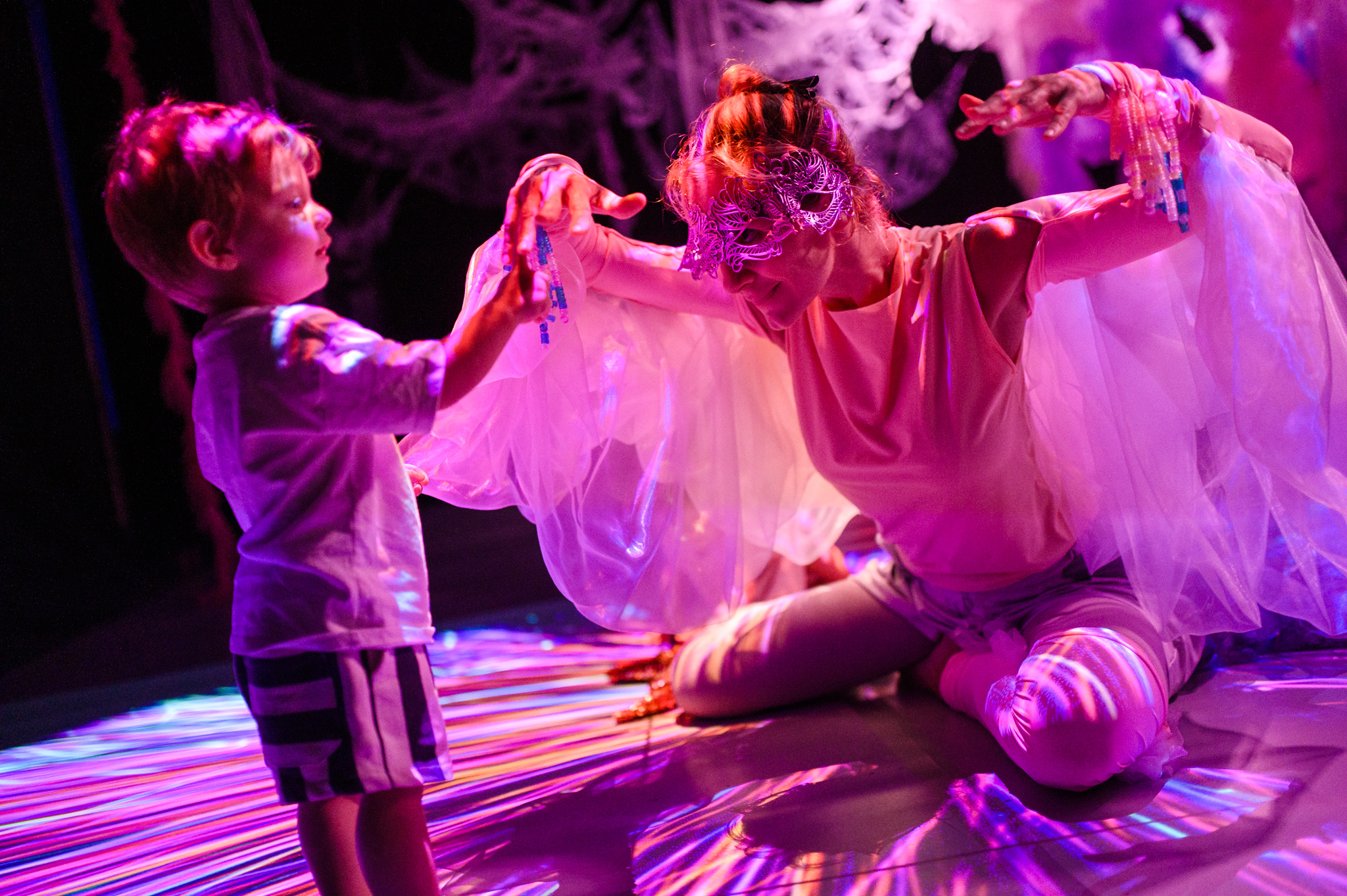 A young child standing and touching the performers hands. The performer is wearing a mask and is kneeling down with their hands up. They have materials hanging from their shoulders resembling wings. 