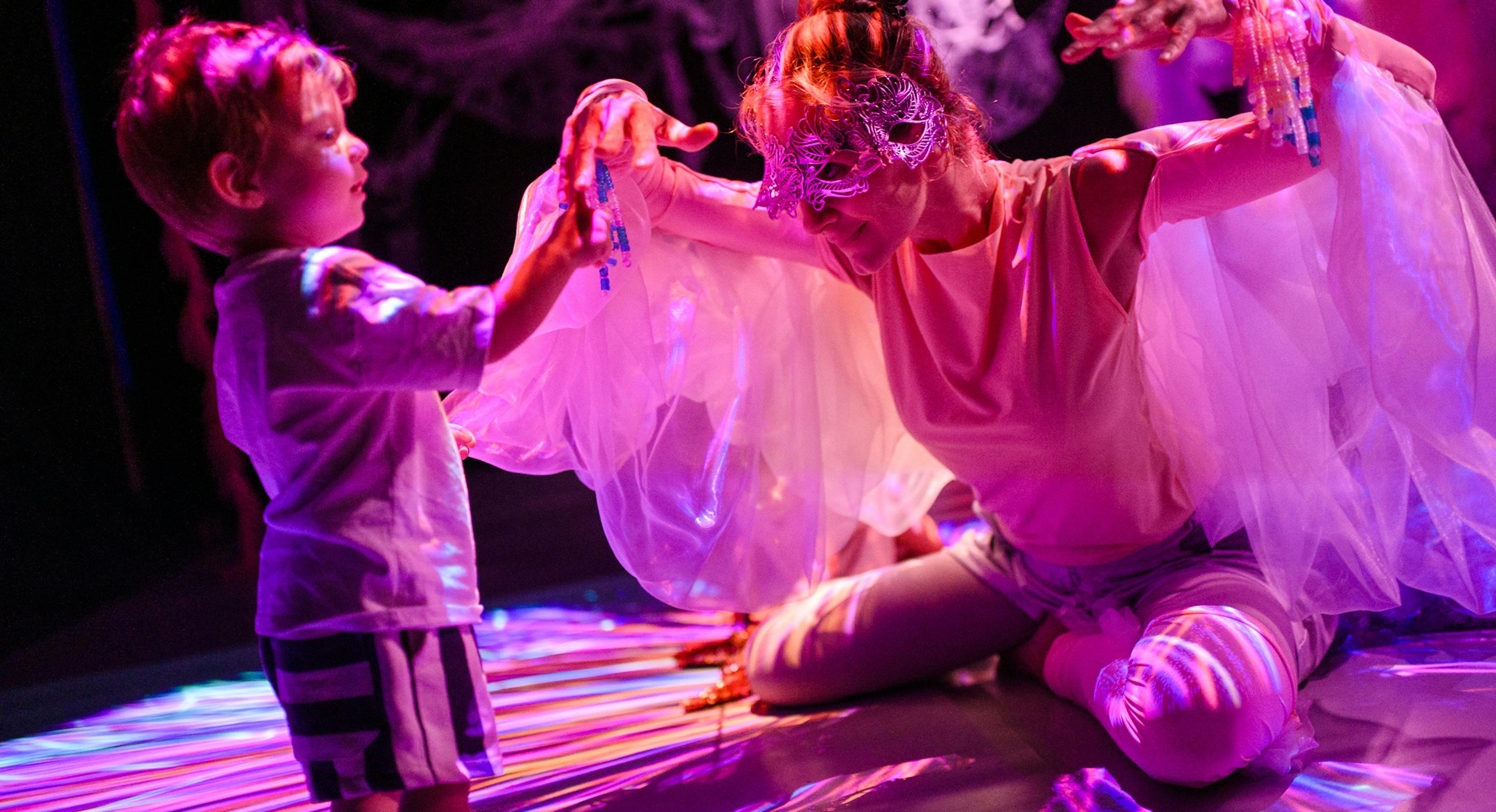 A young child standing and touching the performers hands. The performer is wearing a mask and is kneeling down with their hands up. They have materials hanging from their shoulders resembling wings.