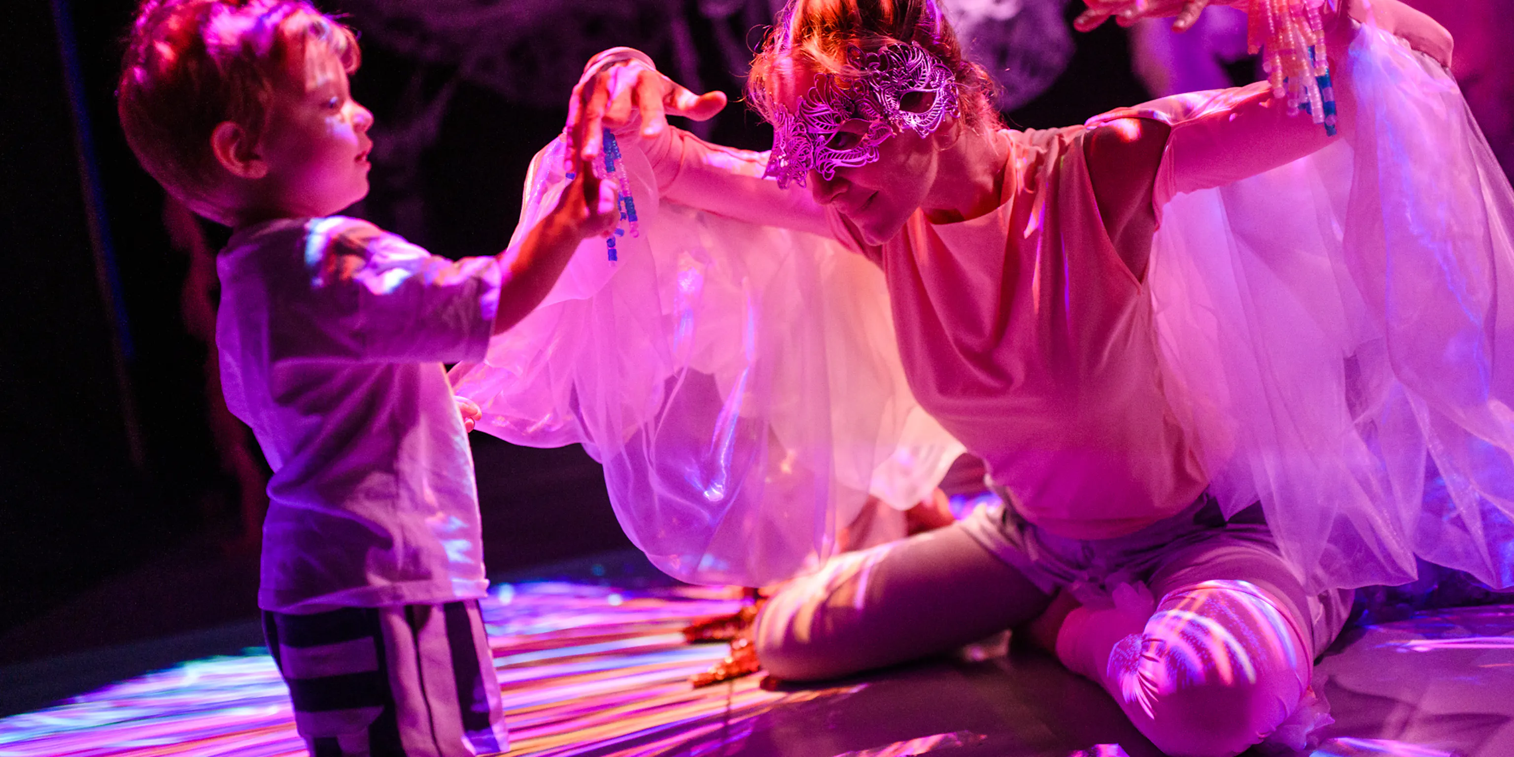 A young child standing and touching the performers hands. The performer is wearing a mask and is kneeling down with their hands up. They have materials hanging from their shoulders resembling wings.