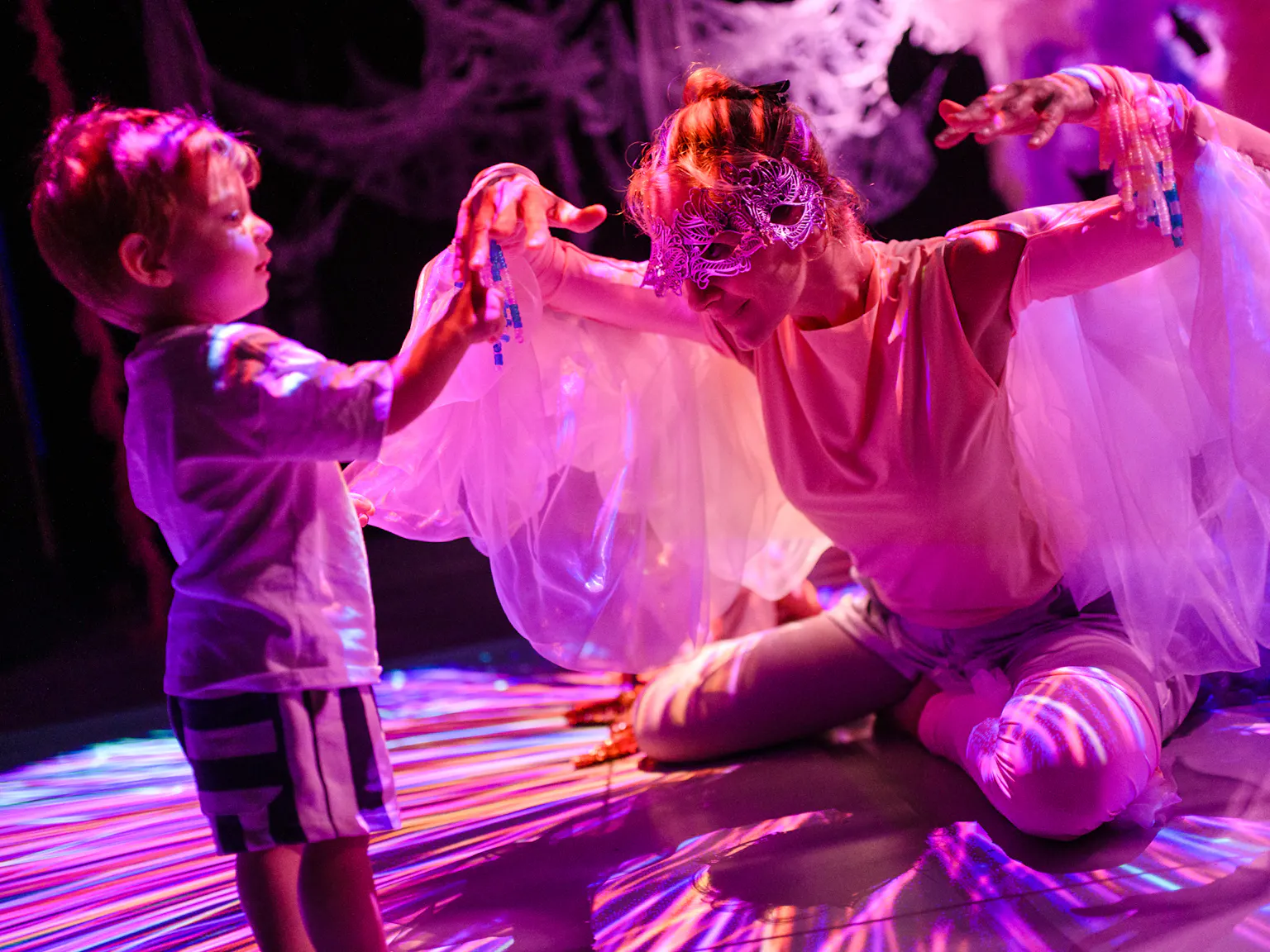 A young child standing and touching the performers hands. The performer is wearing a mask and is kneeling down with their hands up. They have materials hanging from their shoulders resembling wings.