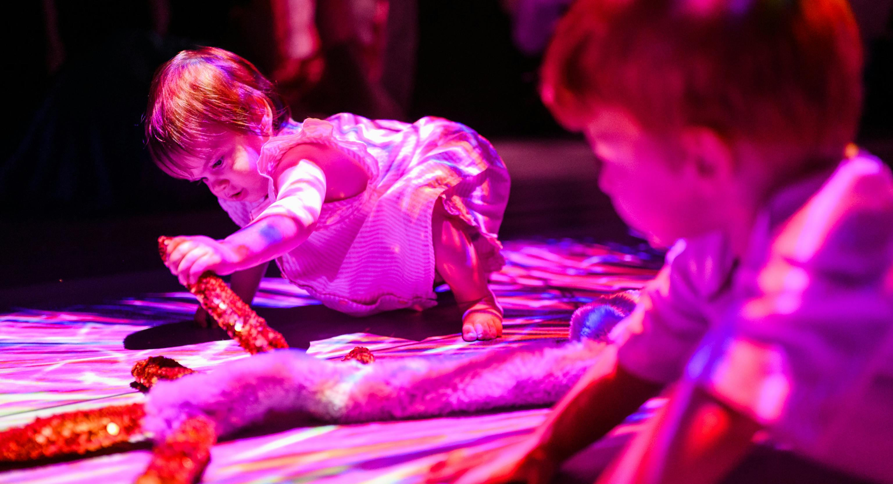 Two children on the floor touching material from the show. One is holding a red sequin piece and the other is watching