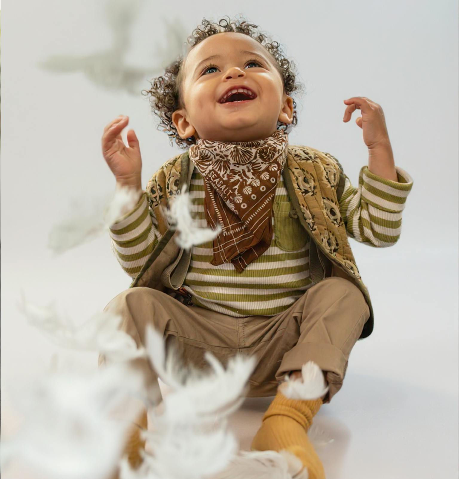 A young child smiling and looking up at feathers falling from the sky