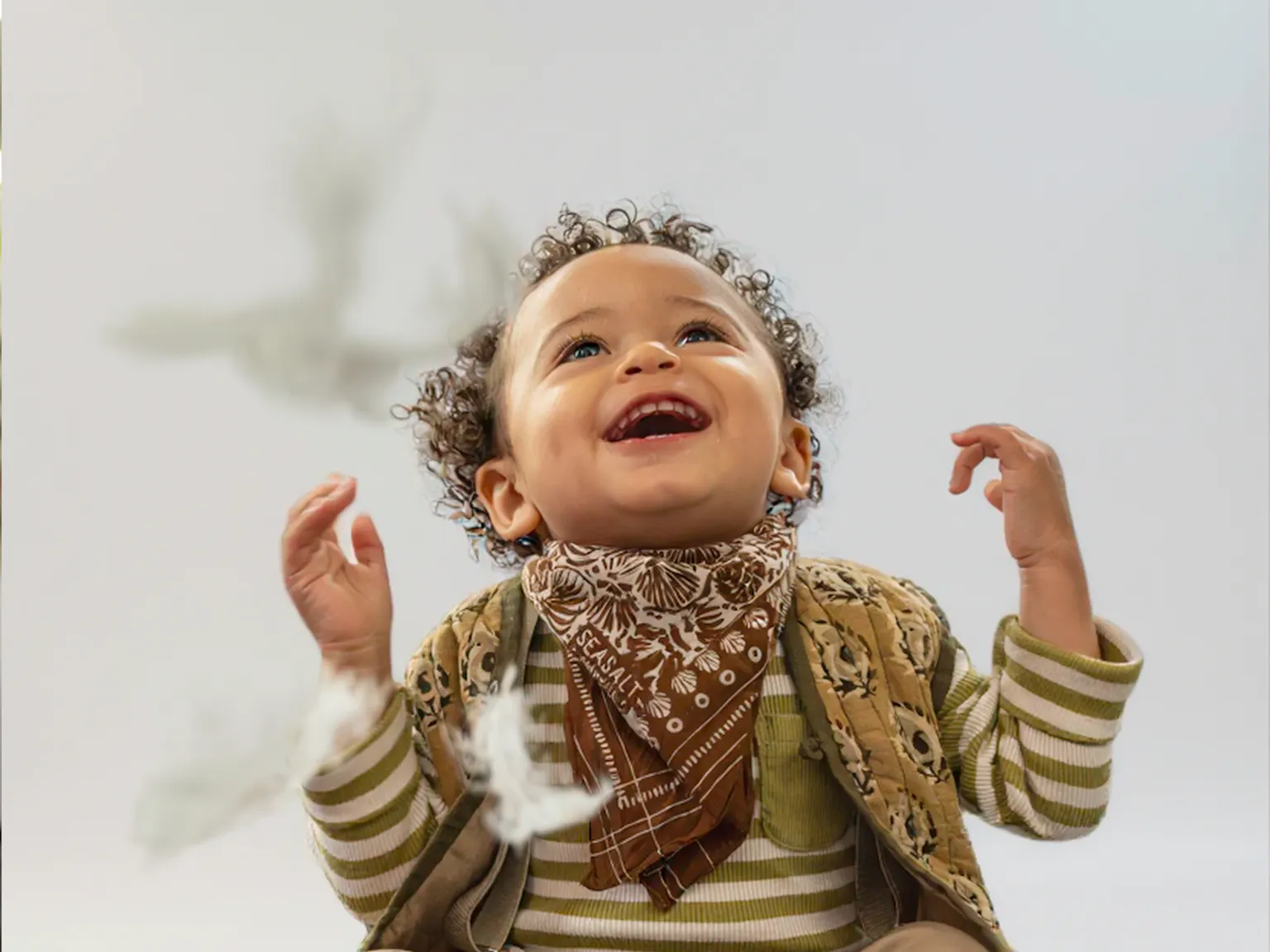 A young child smiling and looking up at feathers falling from the sky