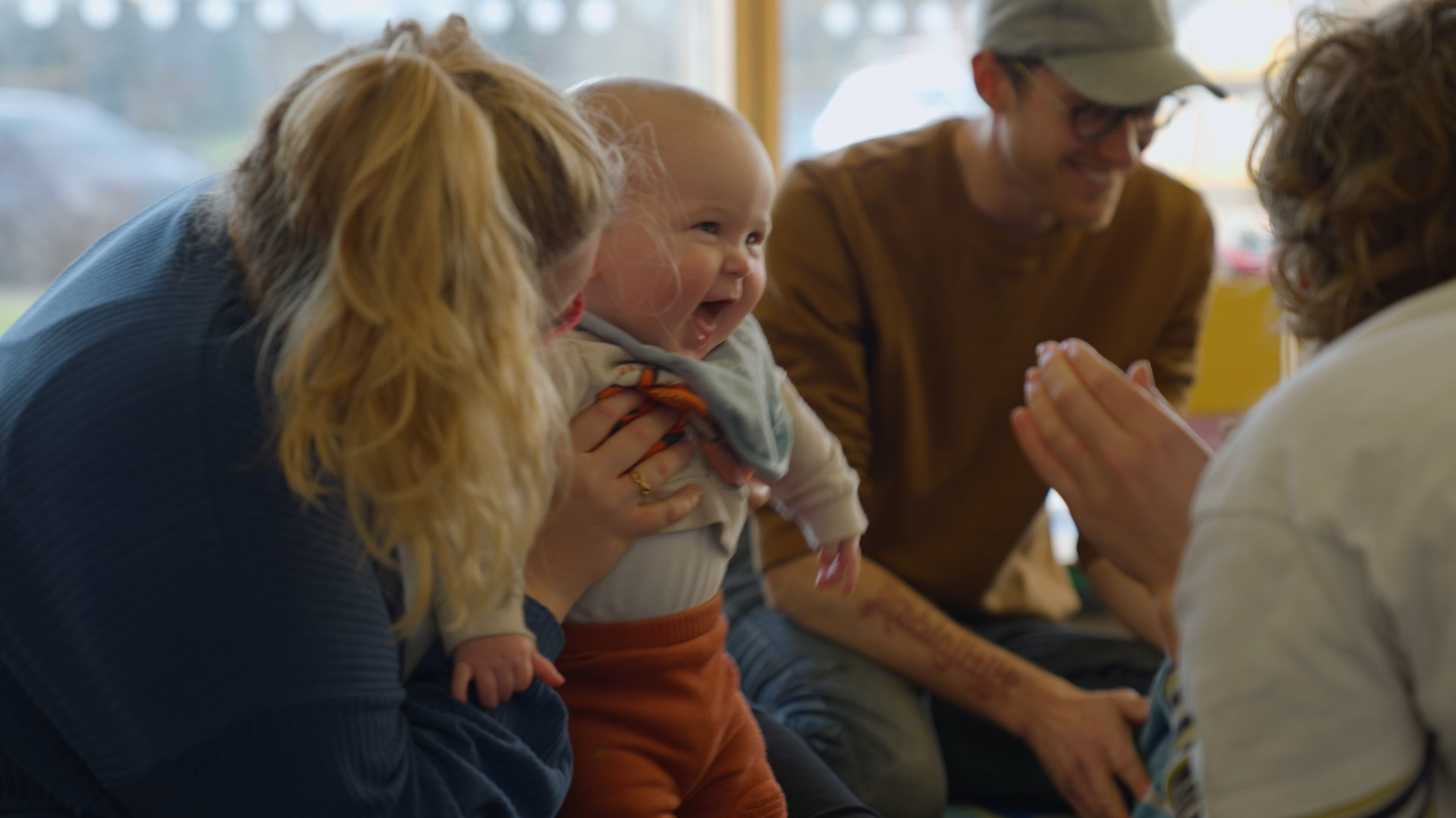 A smiling baby being held up by their parent/carer. The baby is looking towards a playground artist, watching the artist clap. 