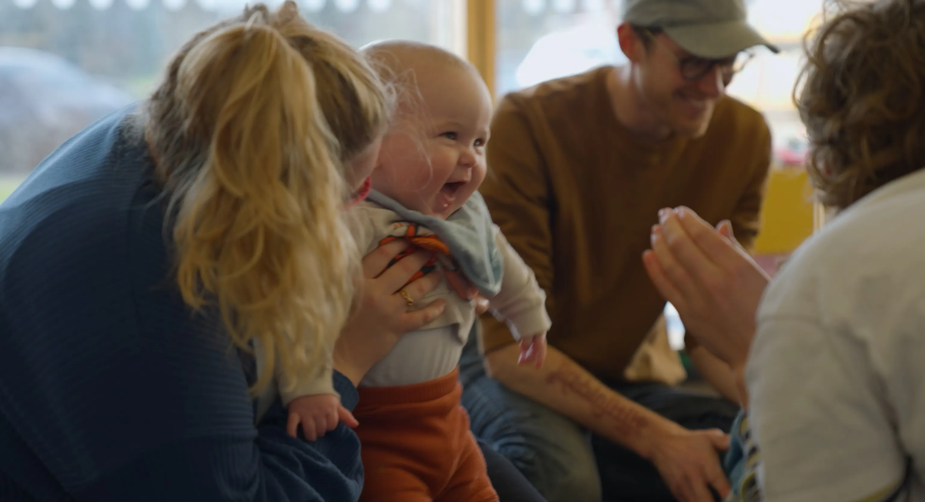 A smiling baby being held up by their parent/carer. The baby is looking towards a playground artist, watching the artist clap.