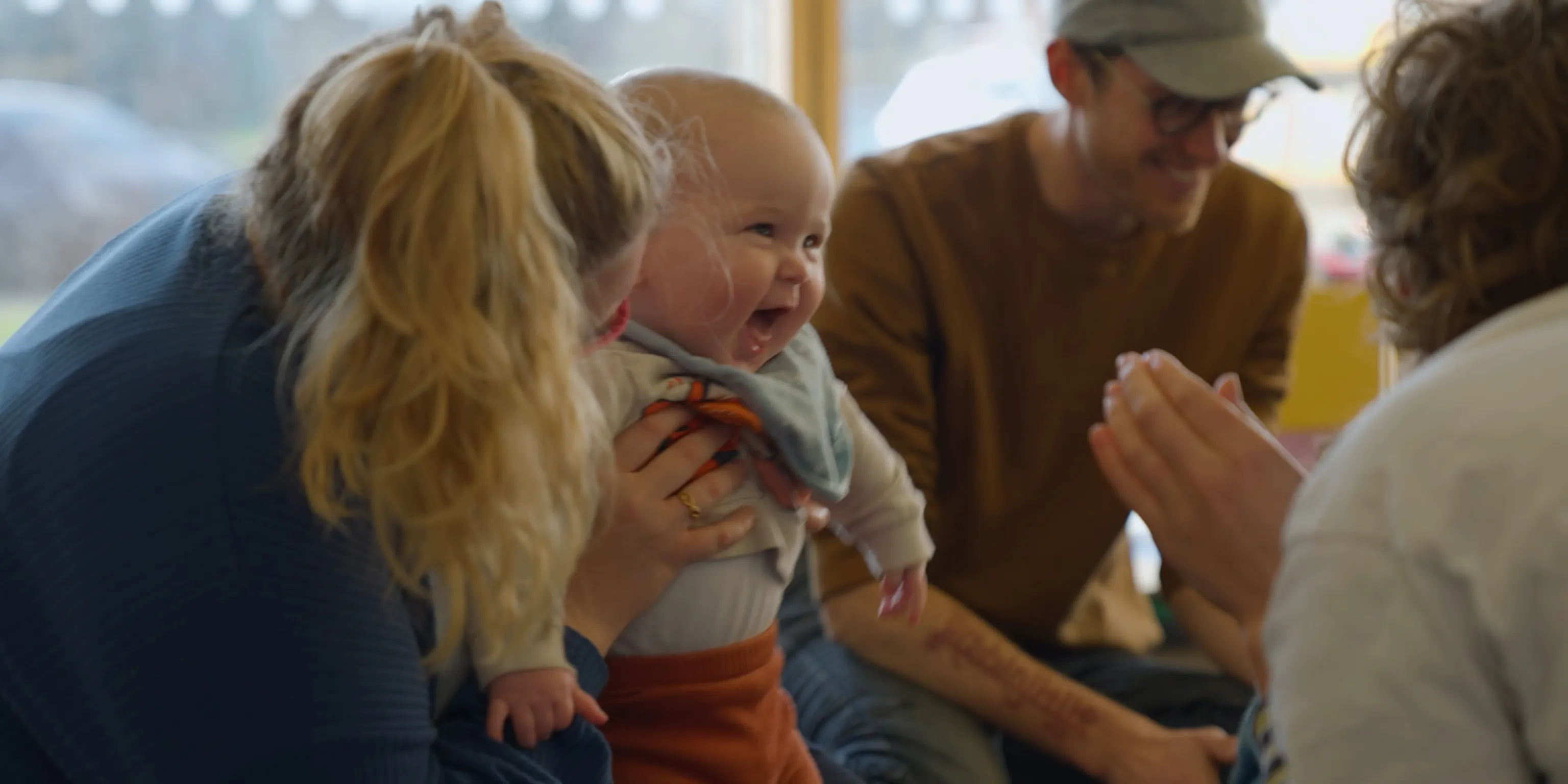 A smiling baby being held up by their parent/carer. The baby is looking towards a playground artist, watching the artist clap.