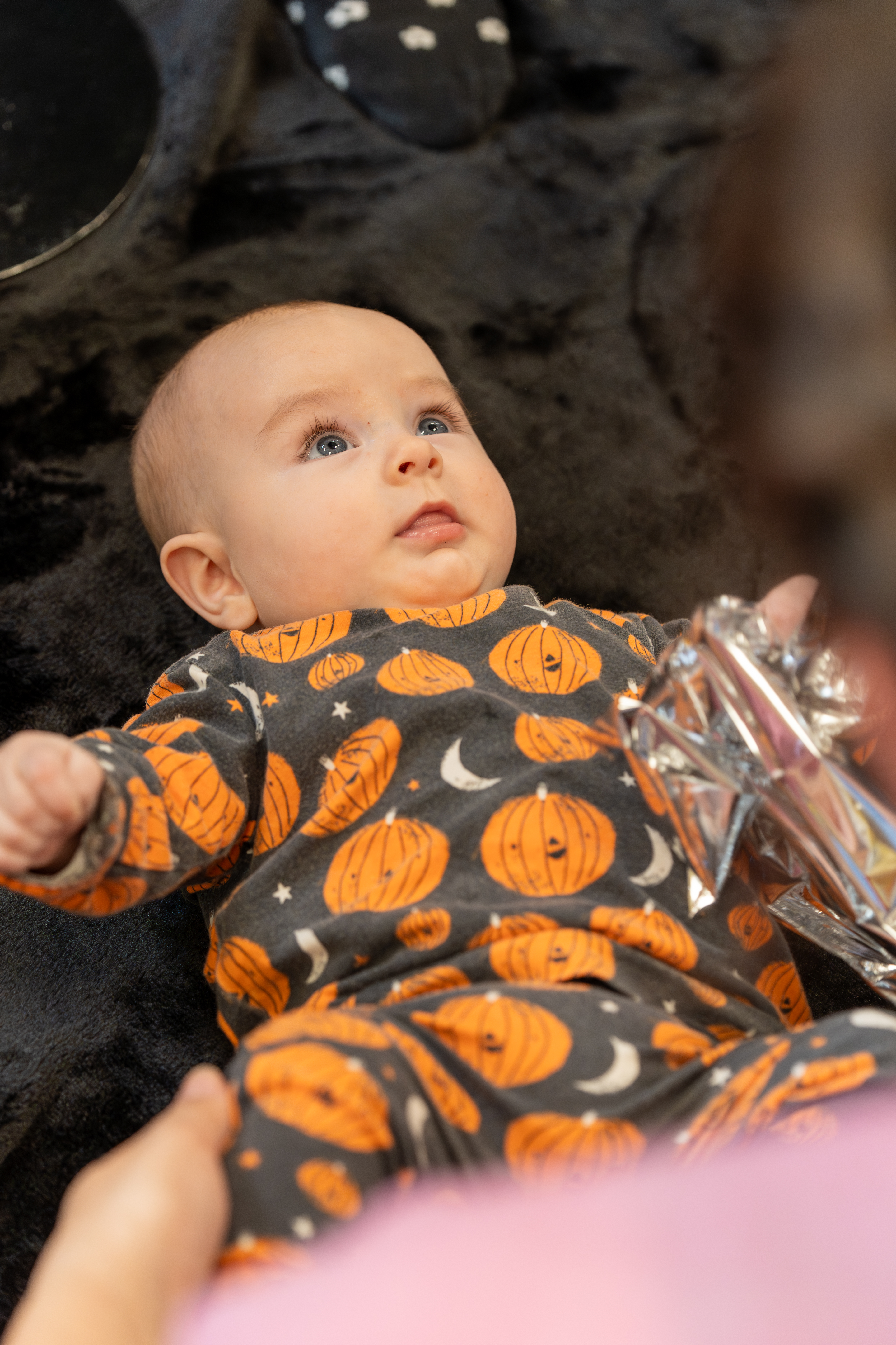 a baby lies on a textured black rug holding a piece of crinkly foil looking up at their parent/carer.
