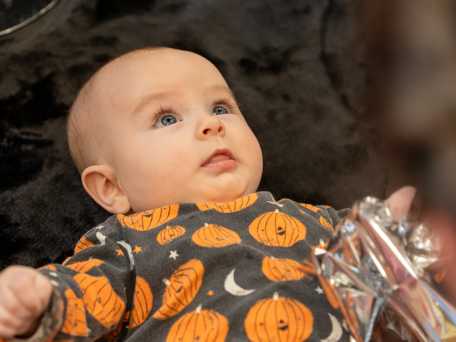 a baby lies on a textured black rug holding a piece of crinkly foil looking up at their parent/carer.