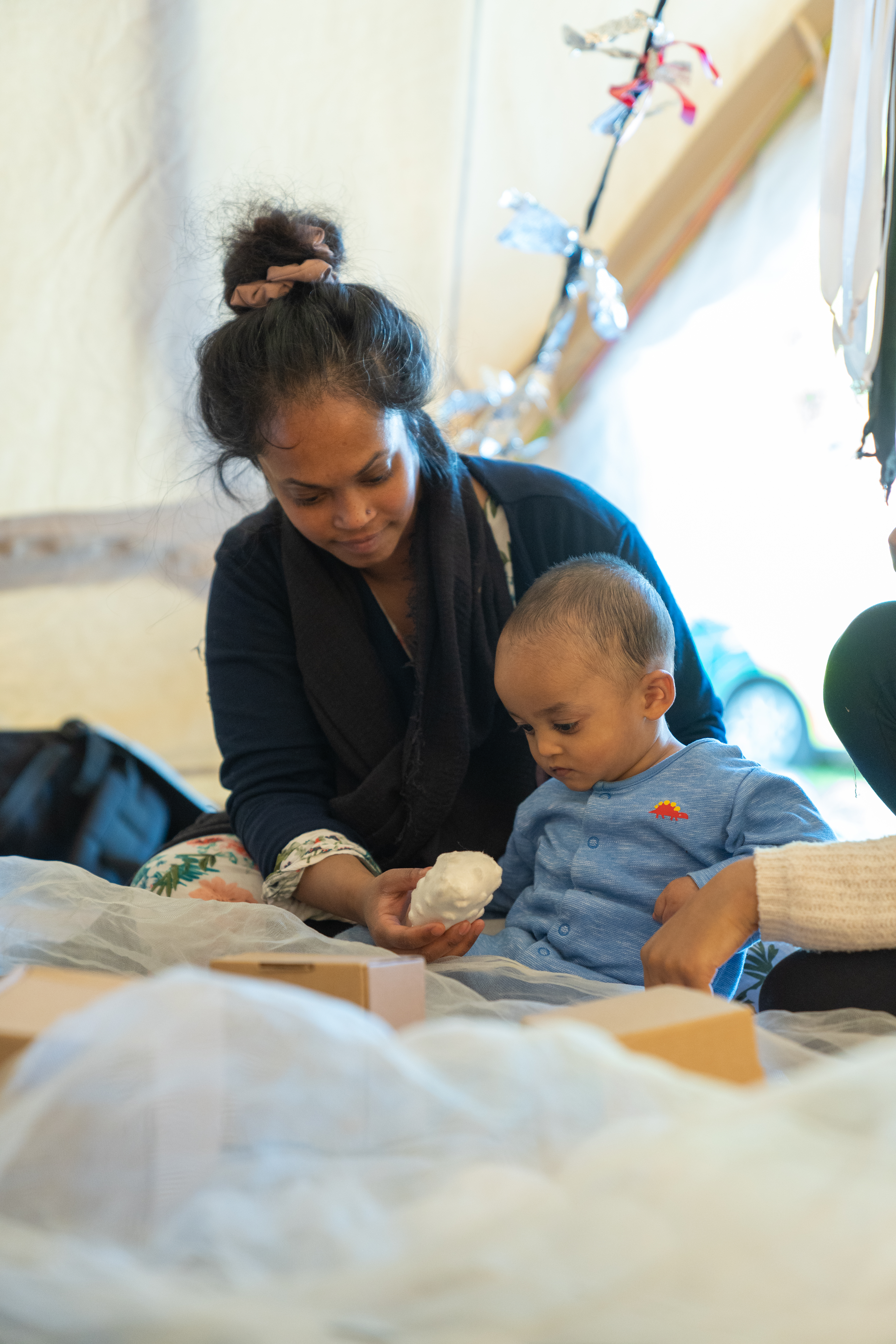 A young child sat surrounded by cardboard boxes and white netting looks down at a ball held by his parent/carer sat behind him.