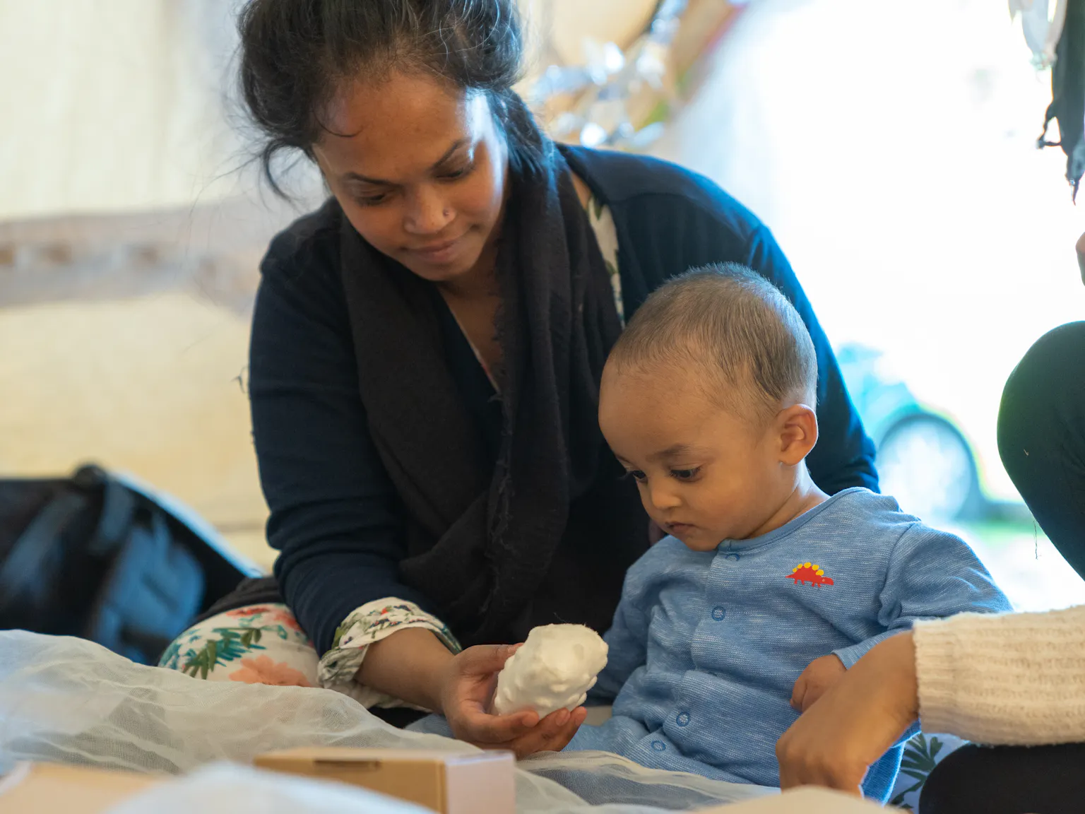 A young child sat surrounded by cardboard boxes and white netting looks down at a ball held by his parent/carer sat behind him.