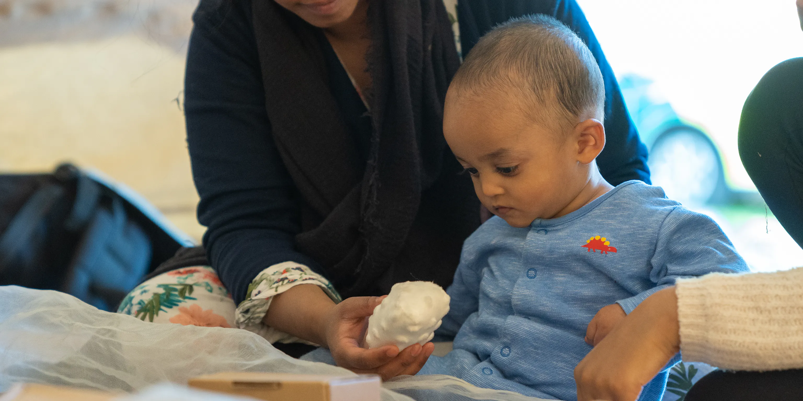 A young child sat surrounded by cardboard boxes and white netting looks down at a ball held by his parent/carer sat behind him.
