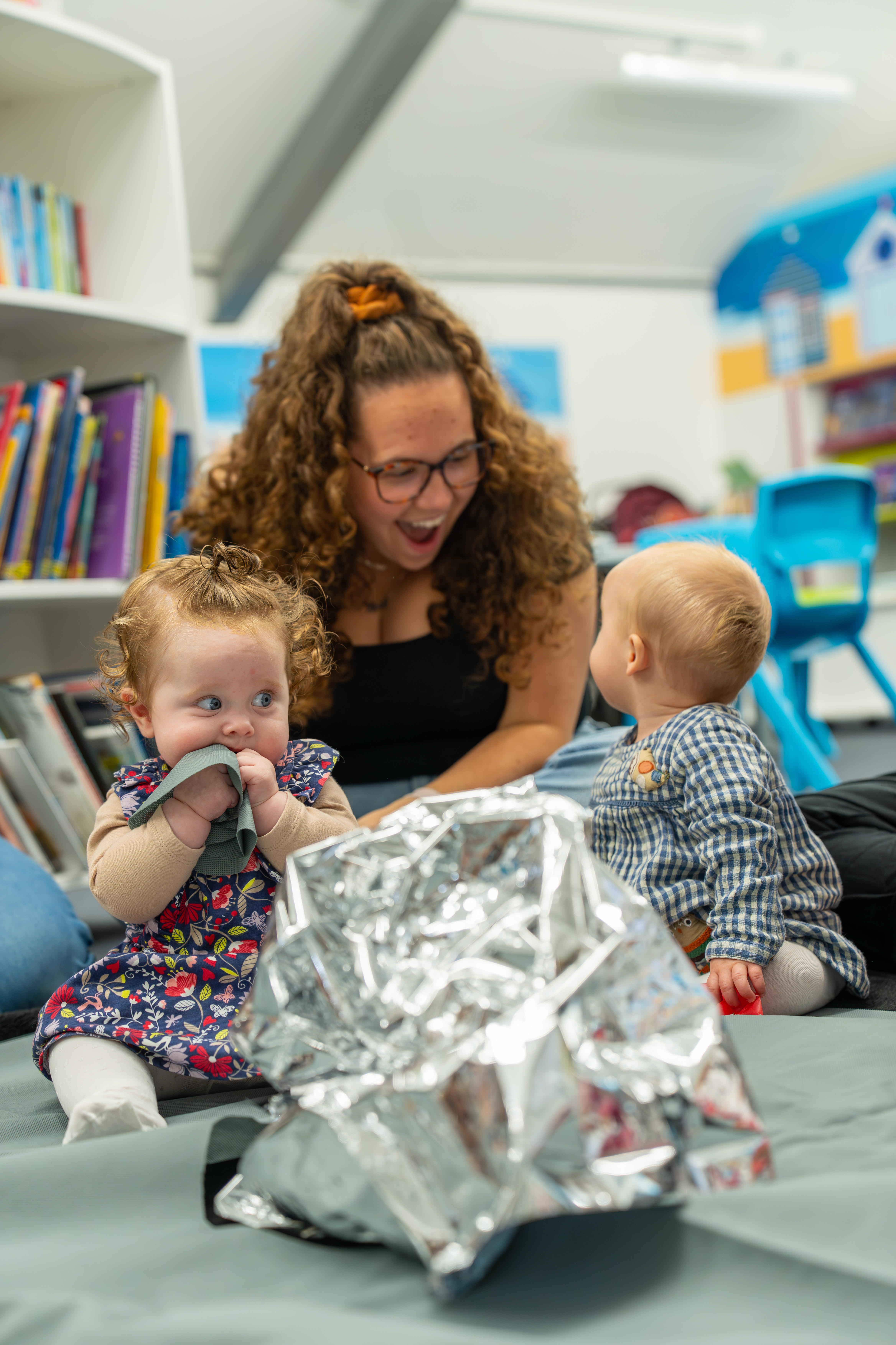 2 babies are sat on top of a grey mat with a large piece of tin foil in front of them. One of them is turned around looking behind them at their parent/carer who is smiling at them. The second baby is sat up scrunching their bib into their mouth.