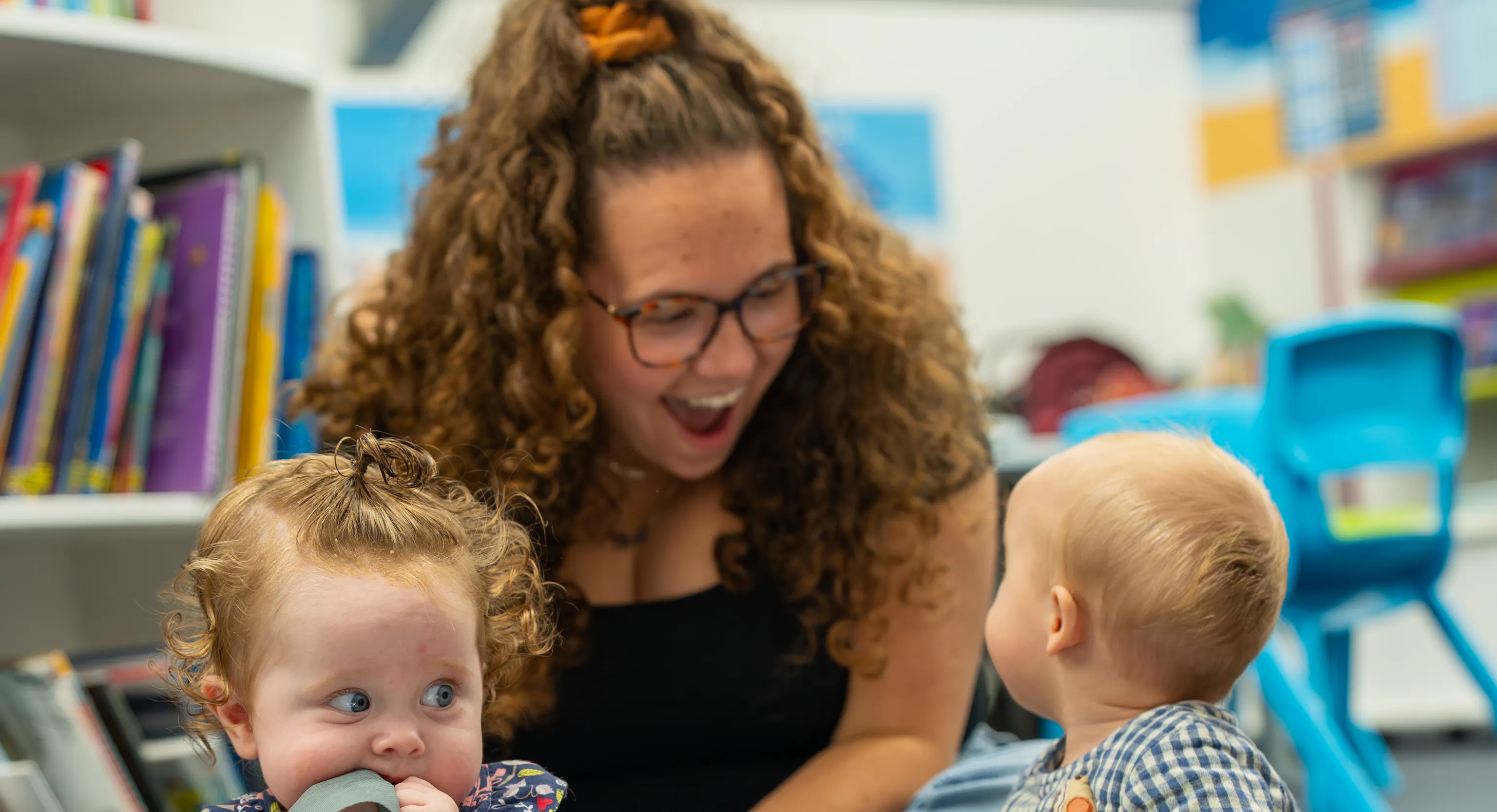 2 babies are sat on top of a grey mat with a large piece of tin foil in front of them. One of them is turned around looking behind them at their parent/carer who is smiling at them. The second baby is sat up scrunching their bib into their mouth.