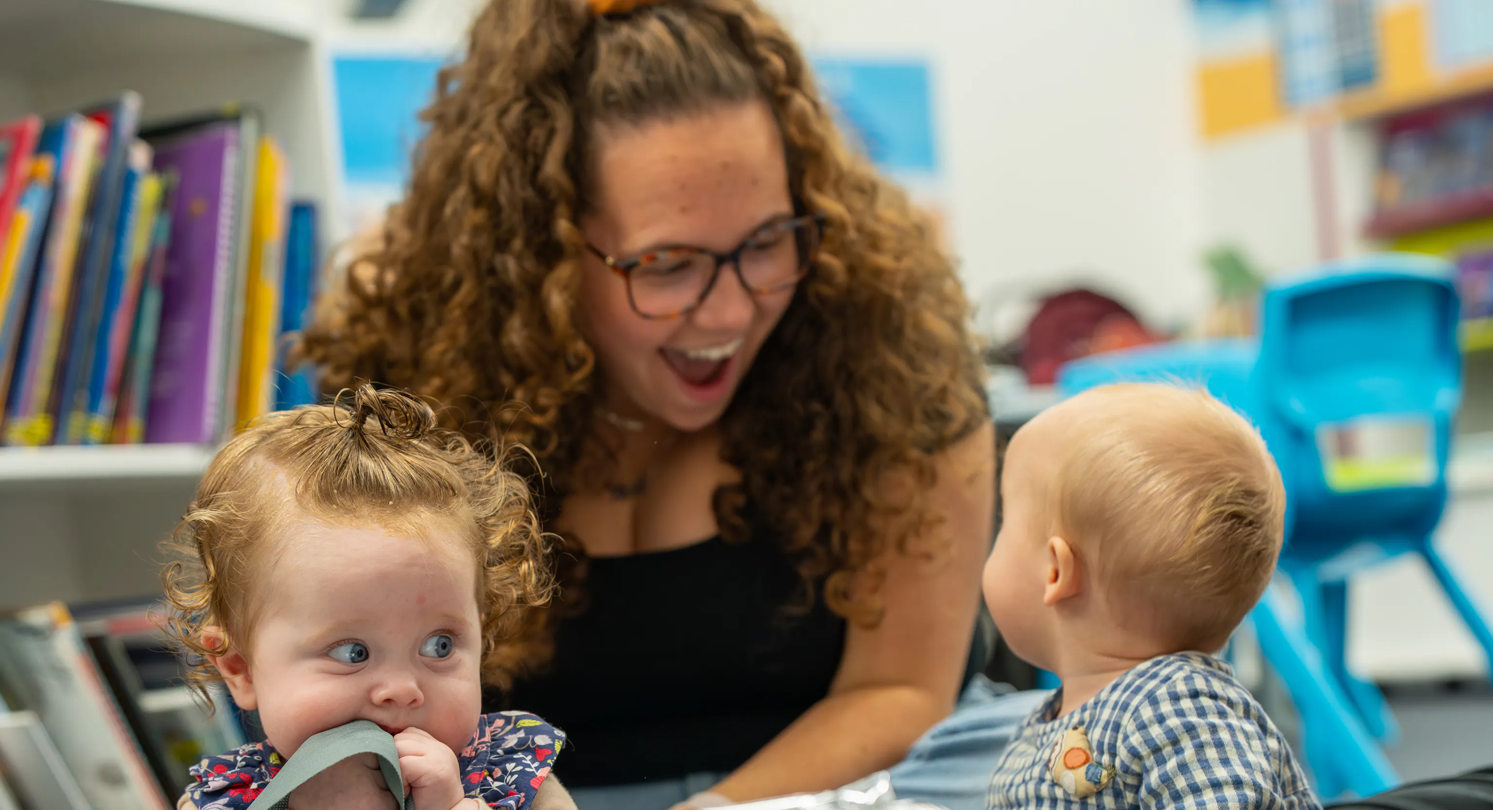 2 babies are sat on top of a grey mat with a large piece of tin foil in front of them. One of them is turned around looking behind them at their parent/carer who is smiling at them. The second baby is sat up scrunching their bib into their mouth.