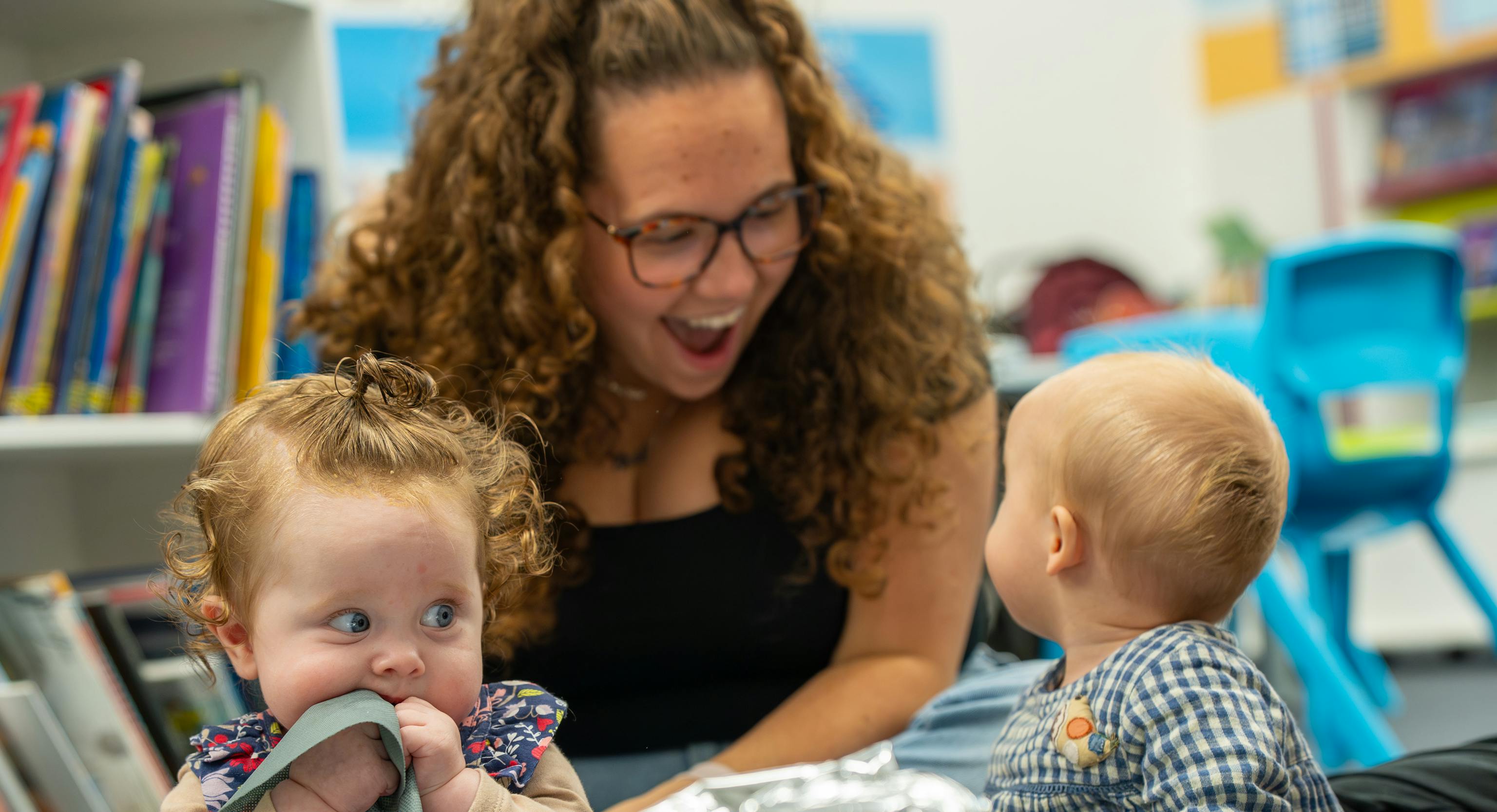 2 babies are sat on top of a grey mat with a large piece of tin foil in front of them. One of them is turned around looking behind them at their parent/carer who is smiling at them. The second baby is sat up scrunching their bib into their mouth.