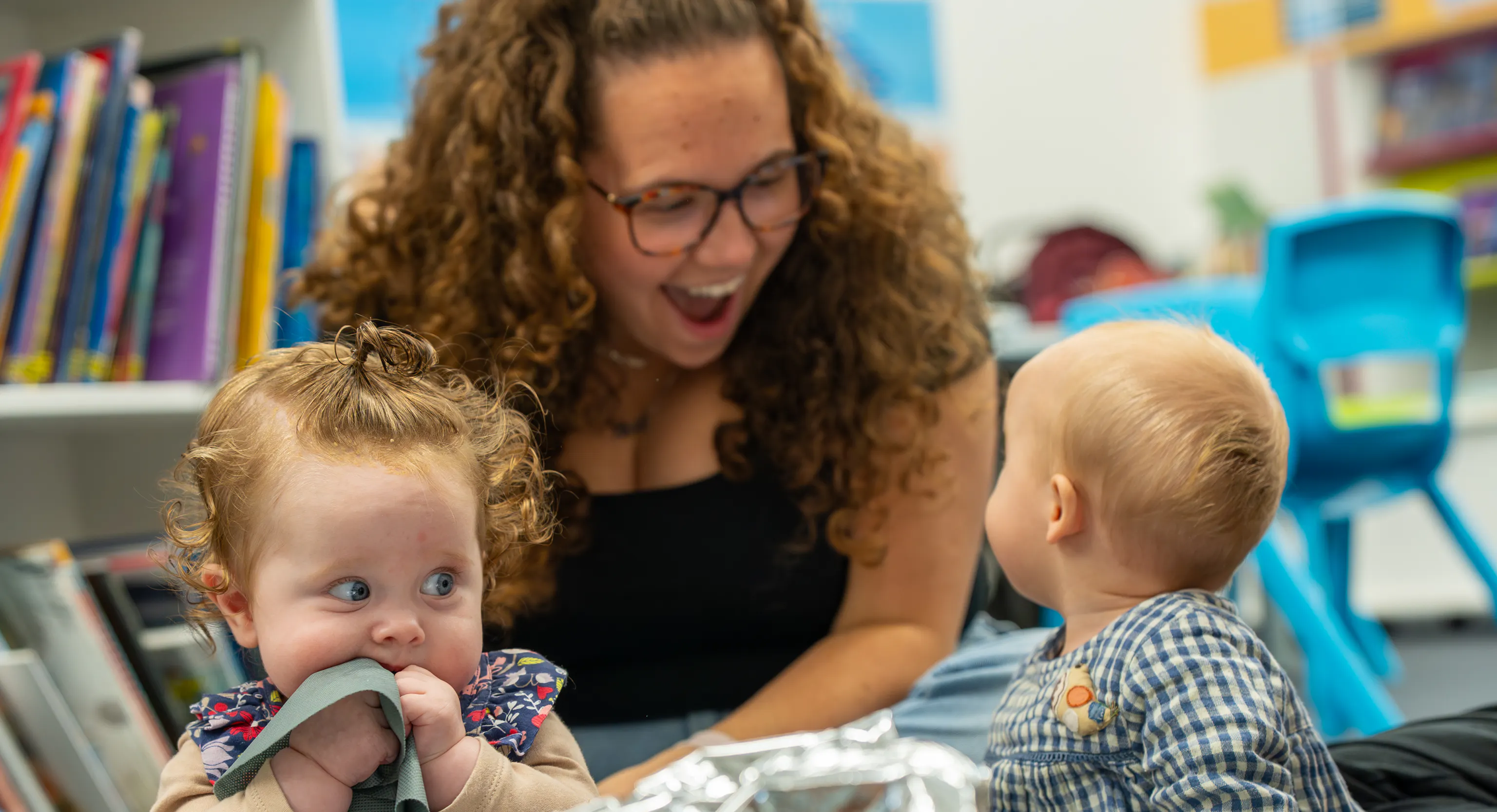 2 babies are sat on top of a grey mat with a large piece of tin foil in front of them. One of them is turned around looking behind them at their parent/carer who is smiling at them. The second baby is sat up scrunching their bib into their mouth.