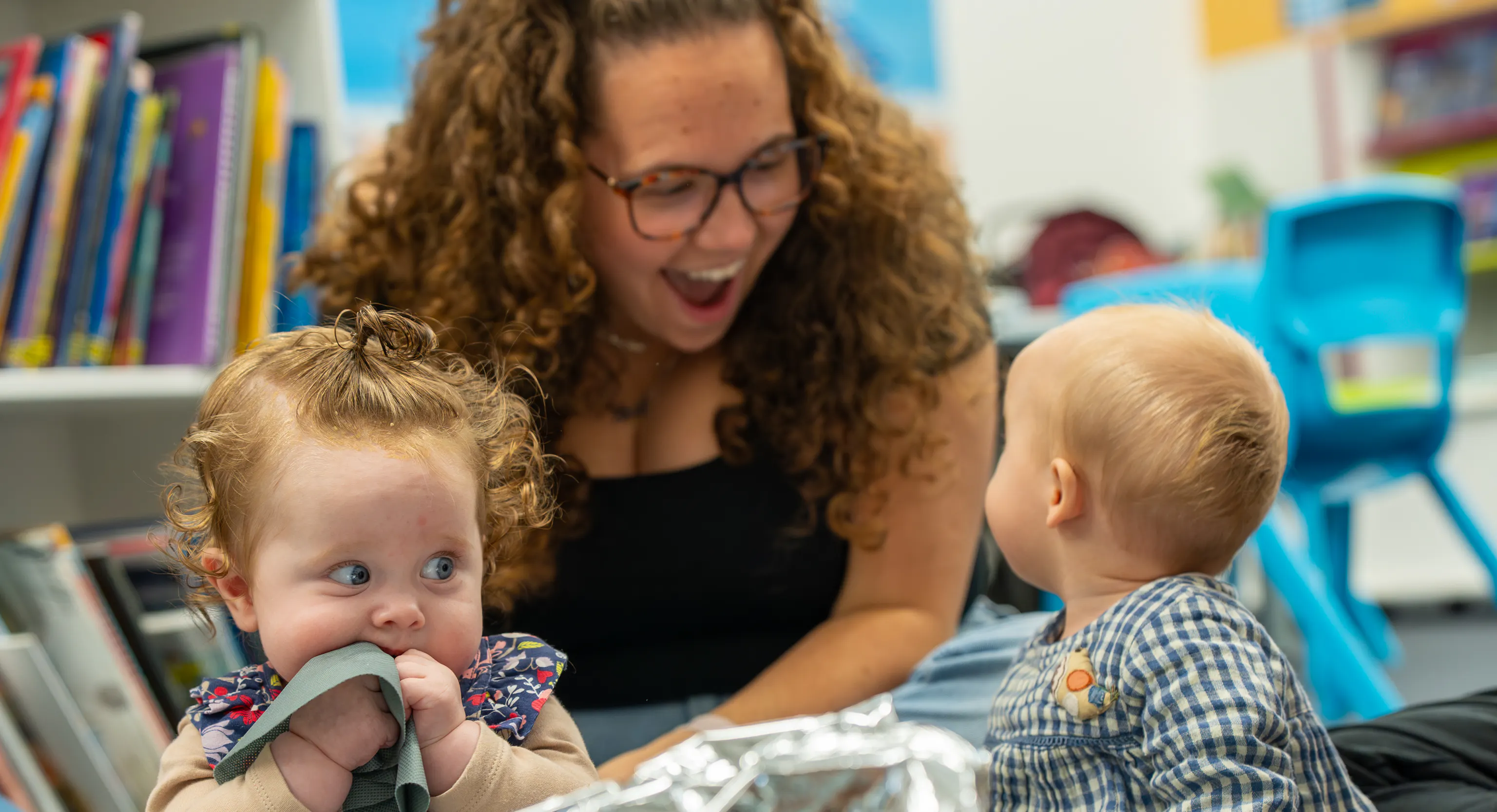 2 babies are sat on top of a grey mat with a large piece of tin foil in front of them. One of them is turned around looking behind them at their parent/carer who is smiling at them. The second baby is sat up scrunching their bib into their mouth.
