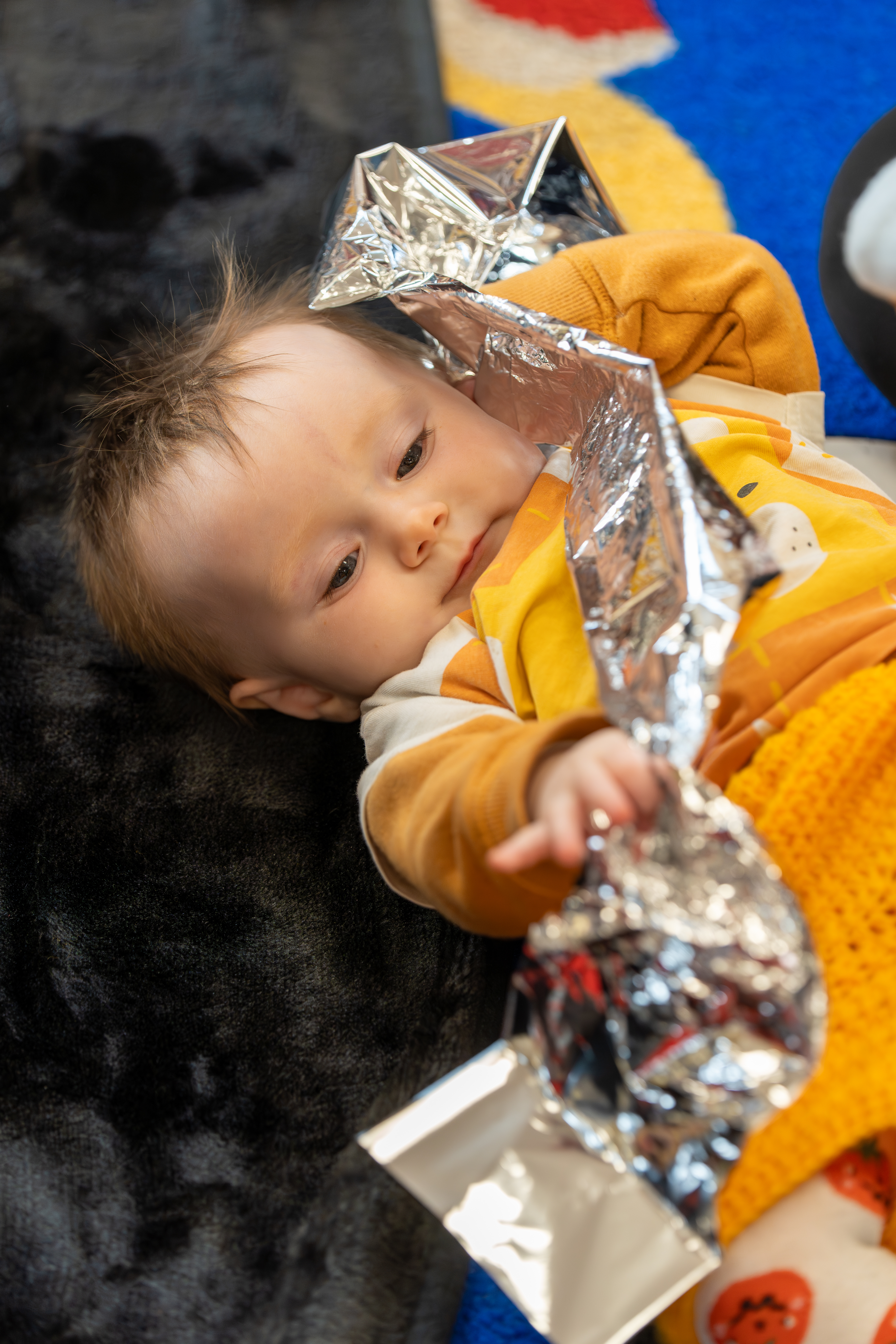 a baby lies on a textured black rug holding a piece of crinkly foil.