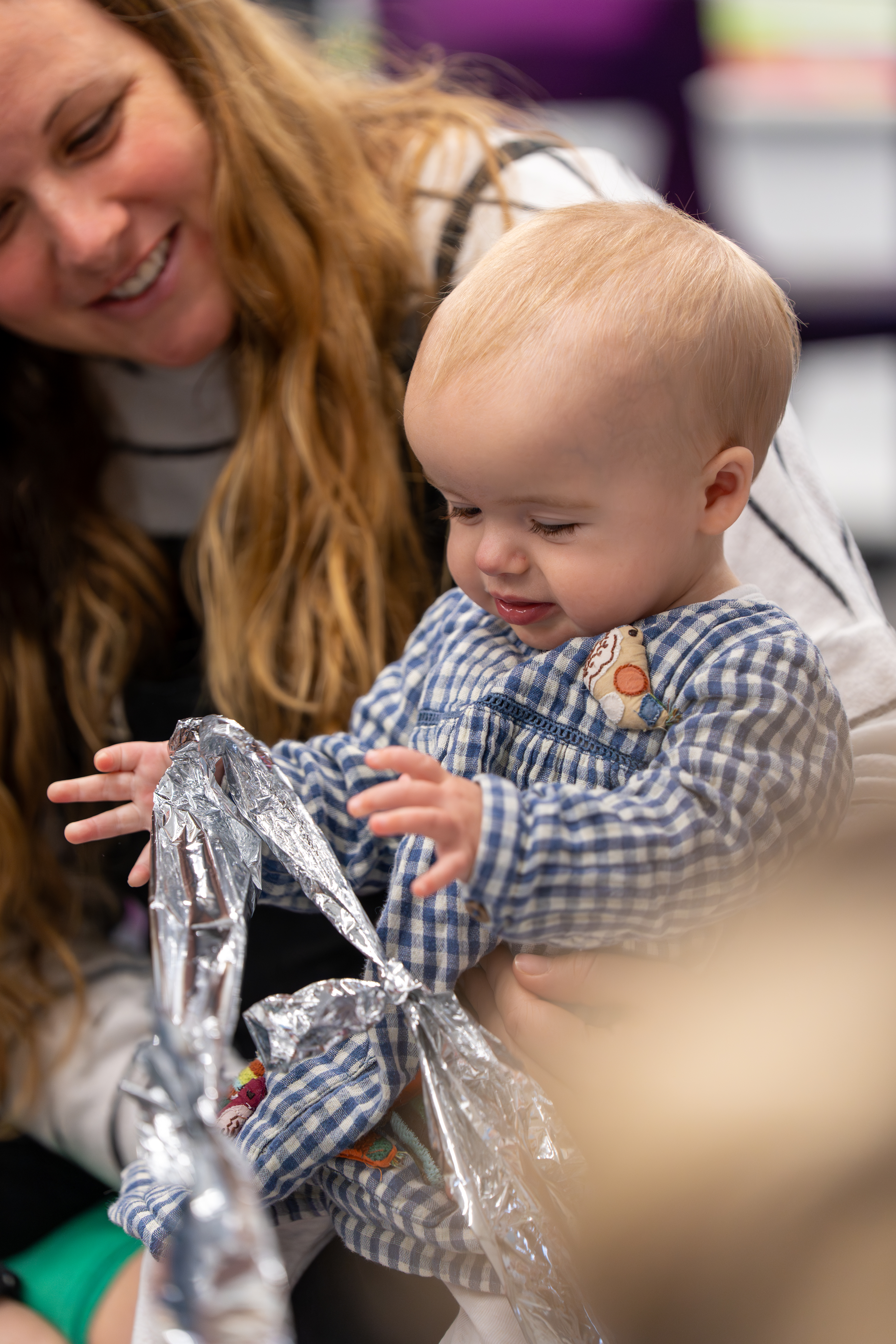 A baby standing up holding a long piece of crinkly foil whilst their parent/carer sits behind them holding the baby up.