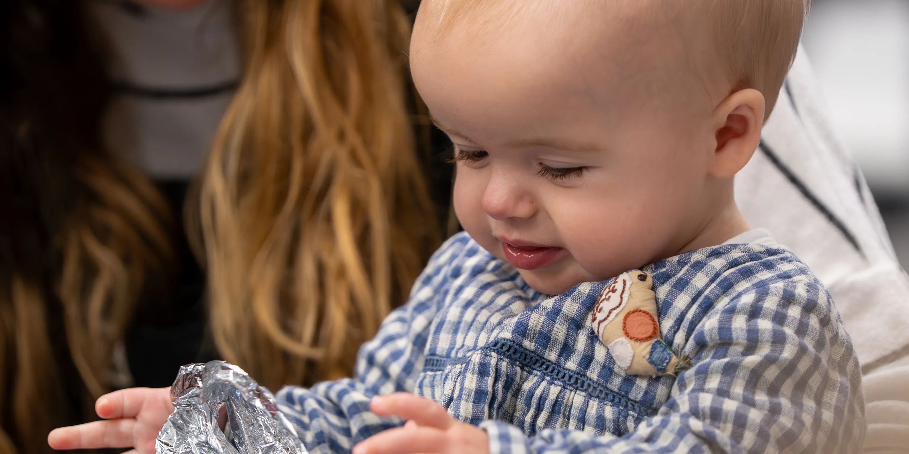 A baby standing up holding a long piece of crinkly foil whilst their parent/carer sits behind them holding the baby up.
