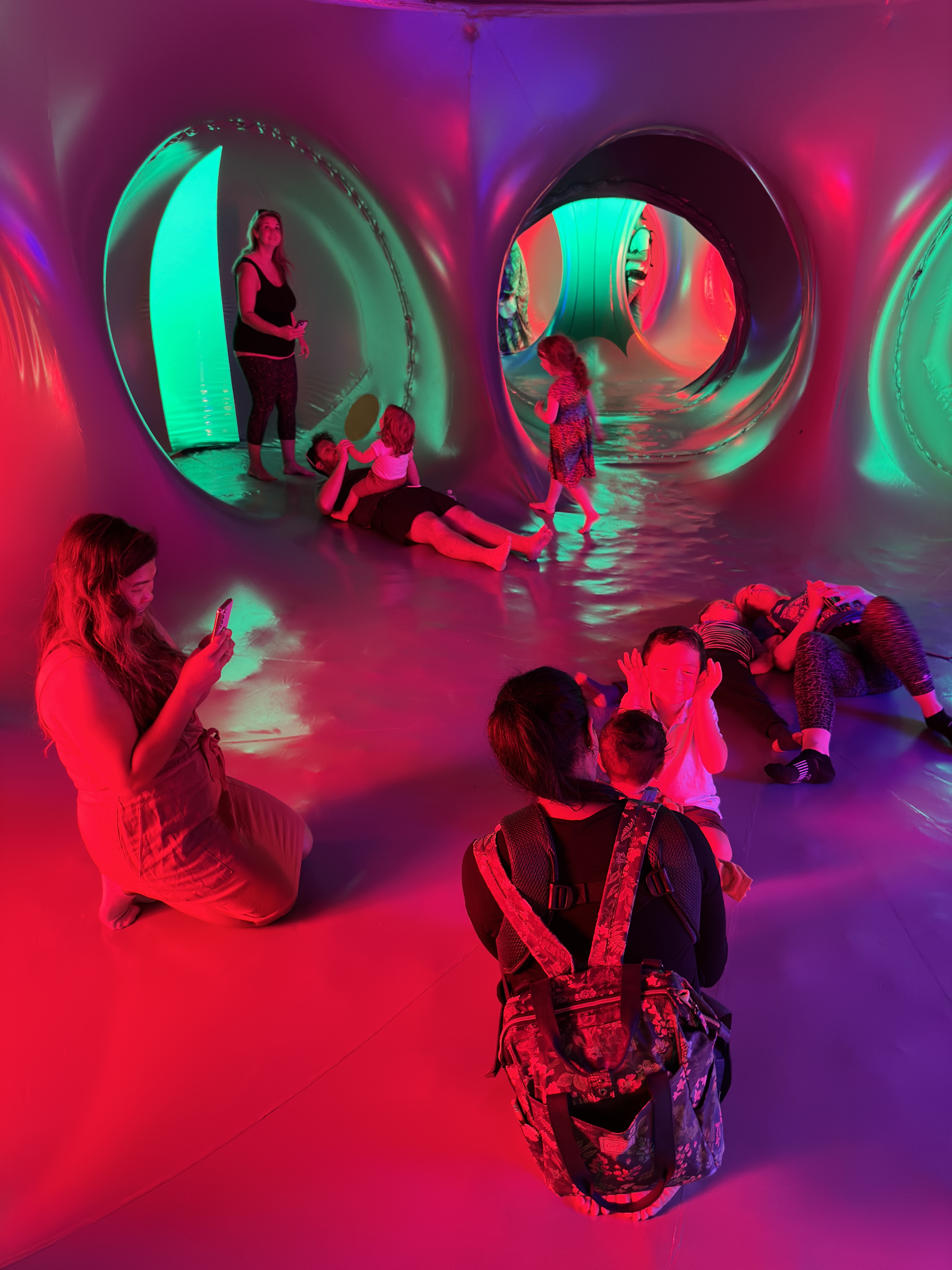 A group of people standing, lying down and kneeling inside of a dome structure with red, green, pink and blue lights