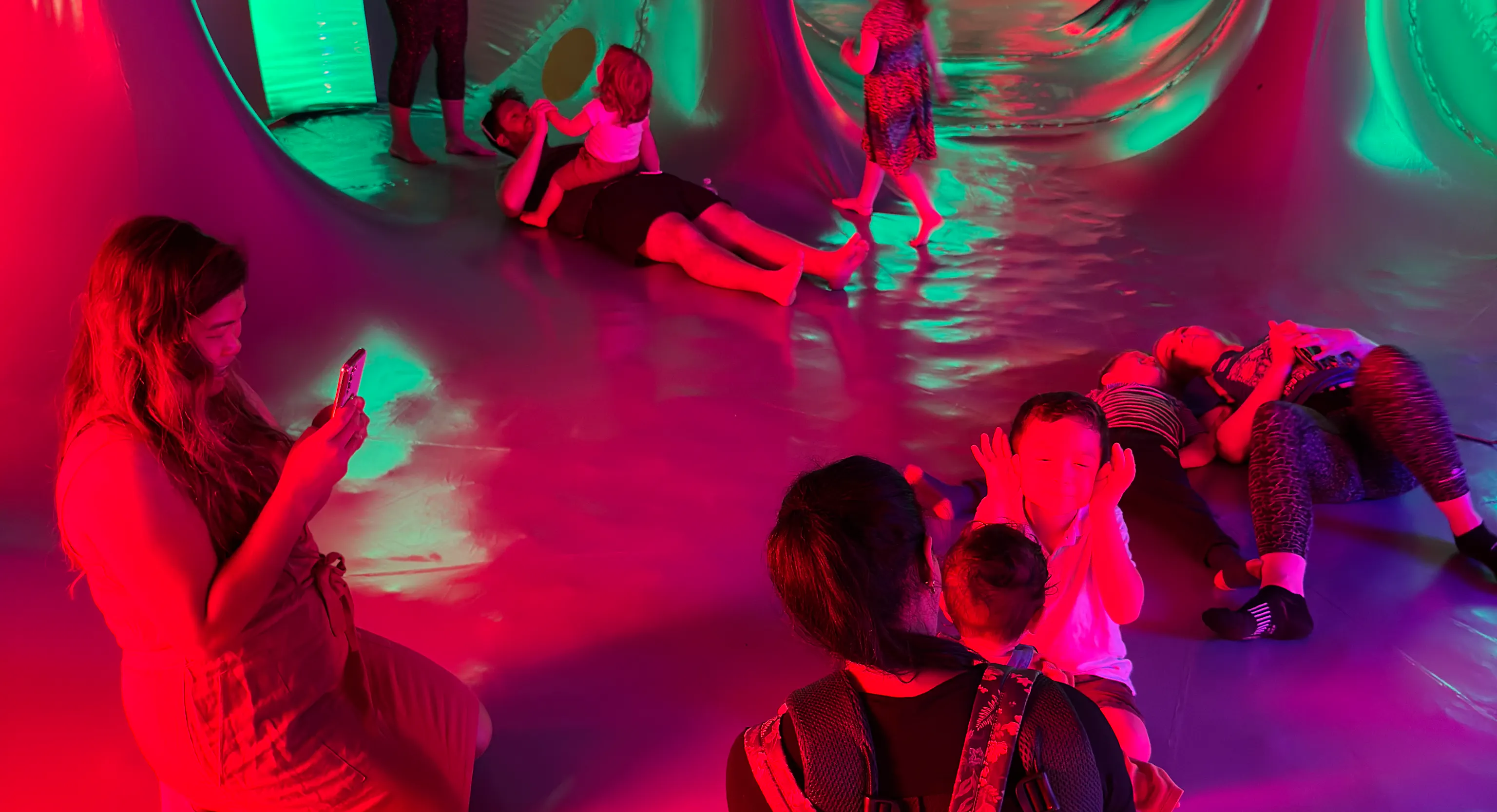 A group of people standing, lying down and kneeling inside of a dome structure with red, green, pink and blue lights