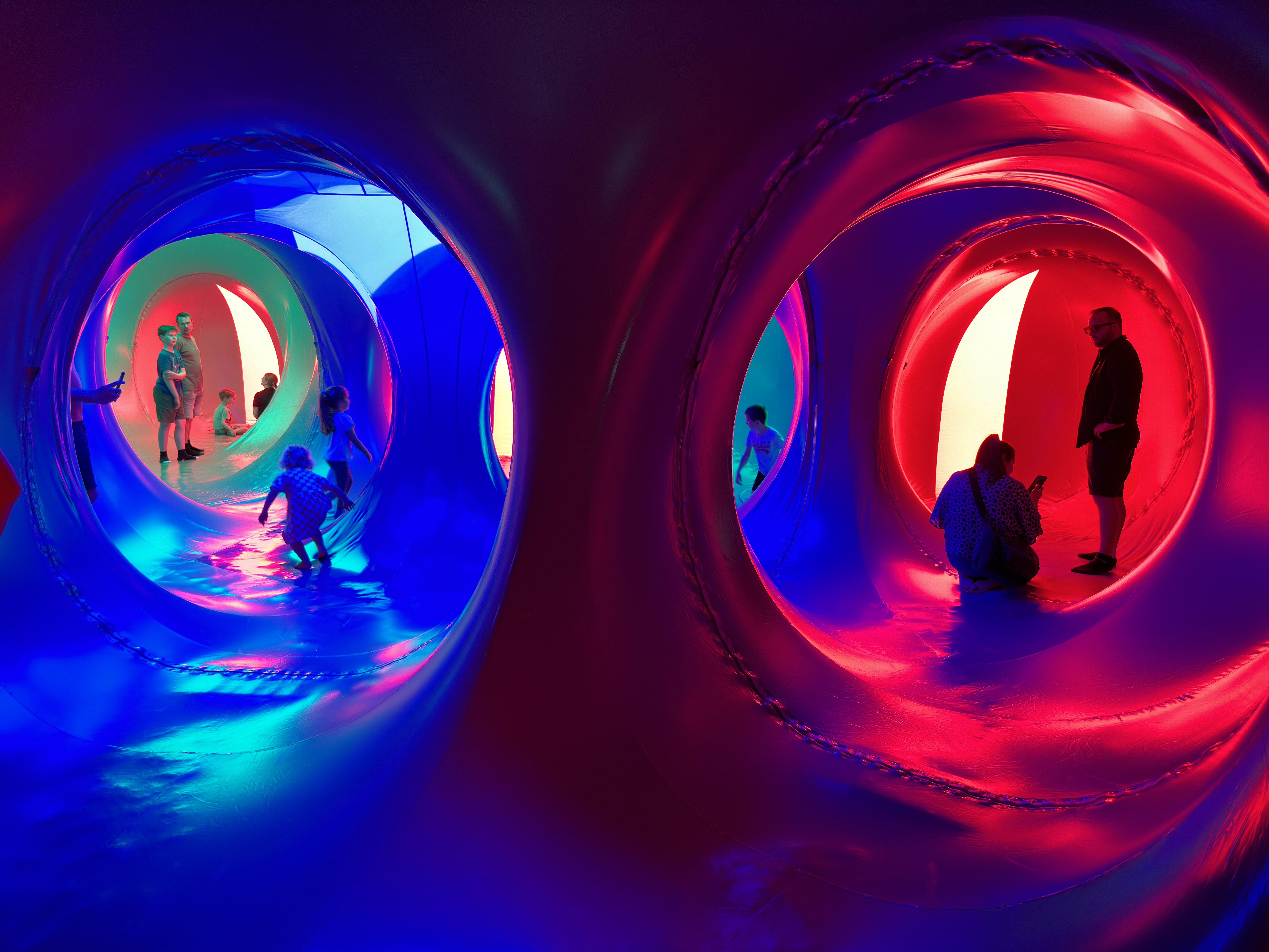 A group of adults and young children stood inside of a dome structure with red, yellow, pink and blue lights