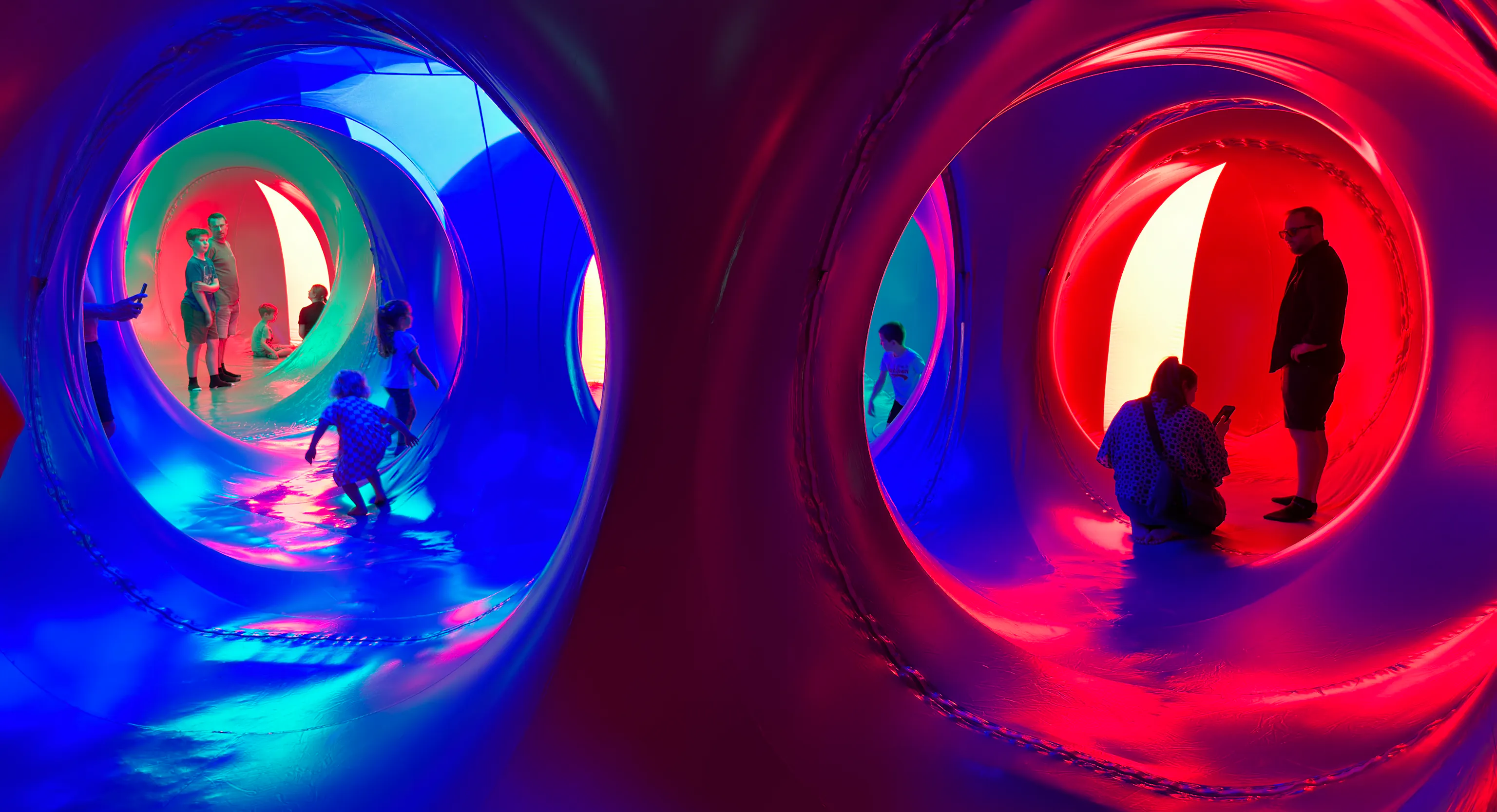 A group of adults and young children stood inside of a dome structure with red, yellow, pink and blue lights