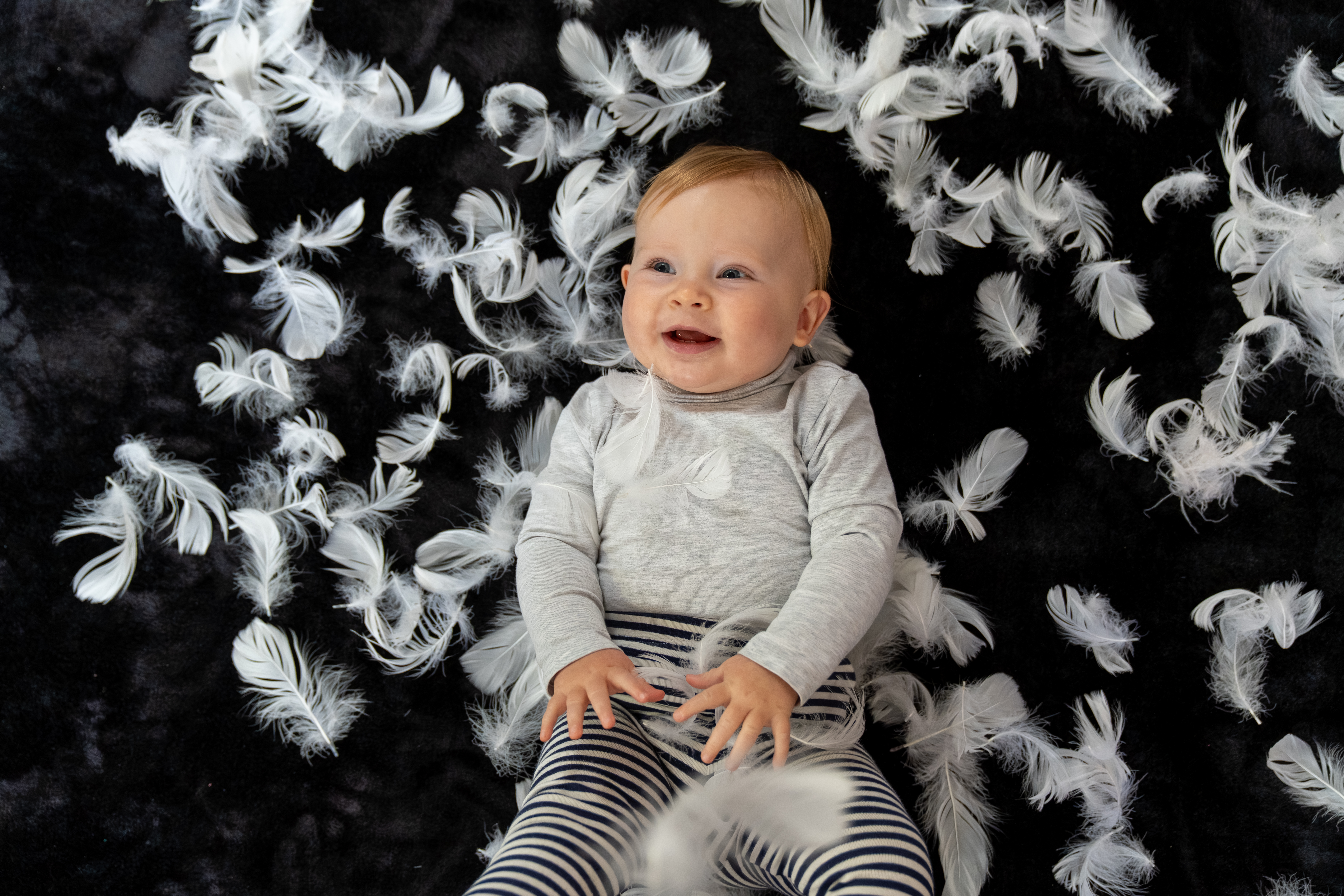 A baby lying on a black mat. She is laughing and is surrounded by feathers 