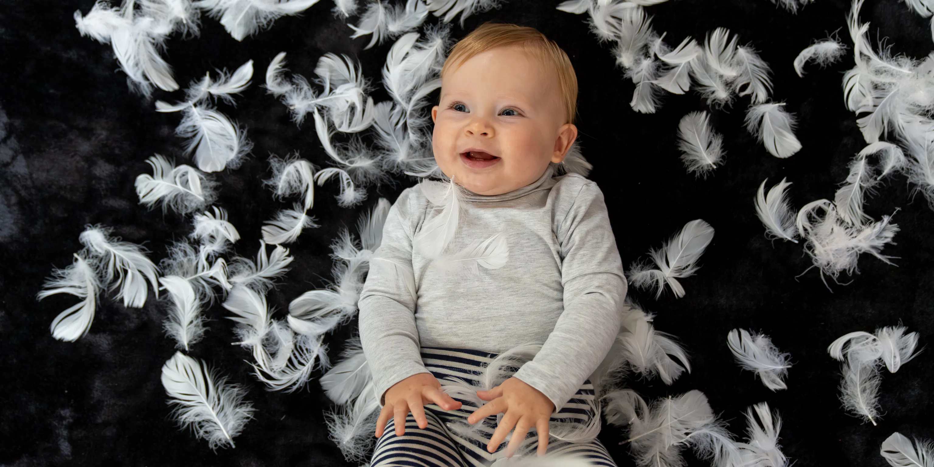 A baby lying on a black mat. She is laughing and is surrounded by feathers