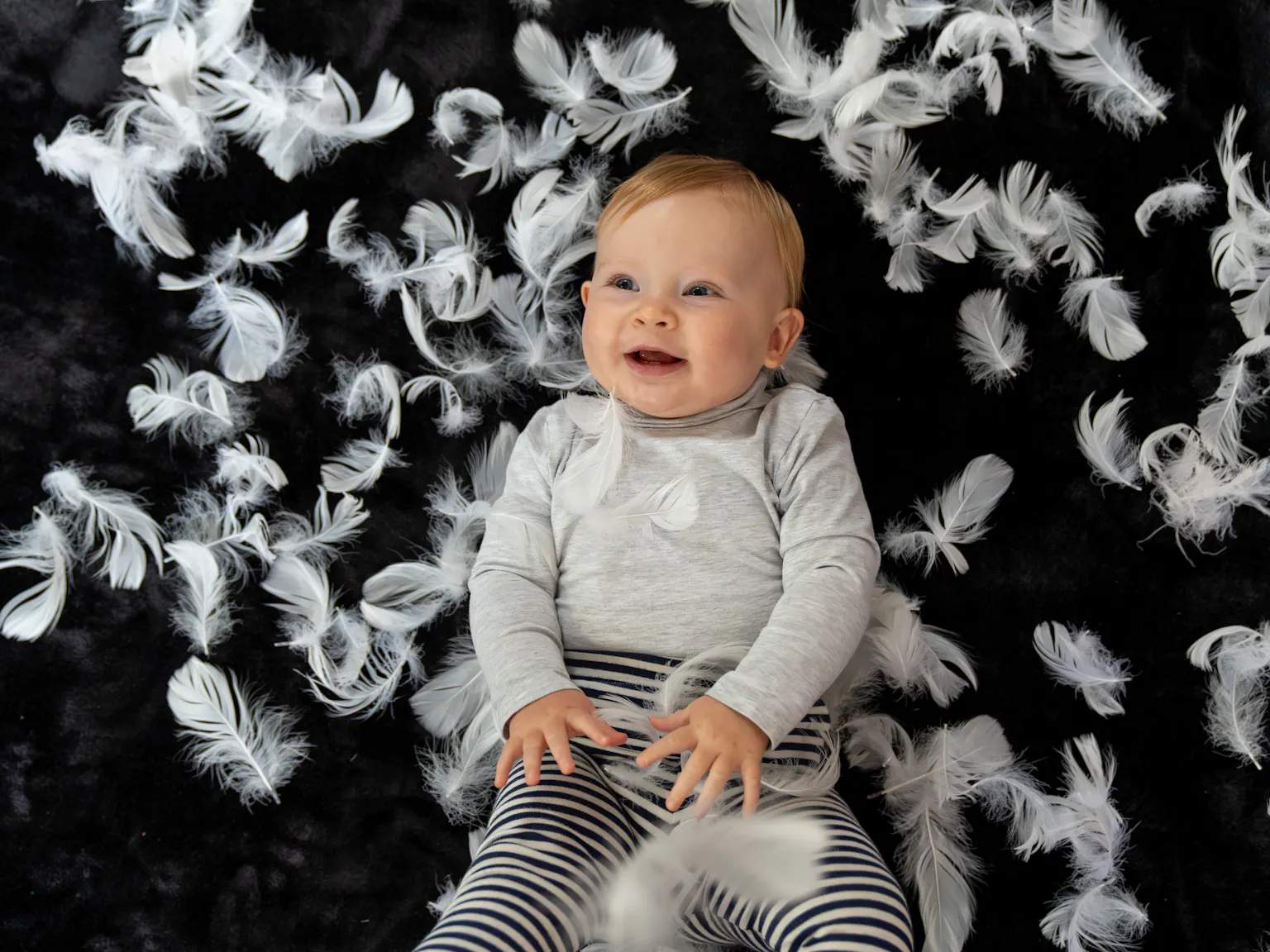 A baby lying on a black mat. She is laughing and is surrounded by feathers