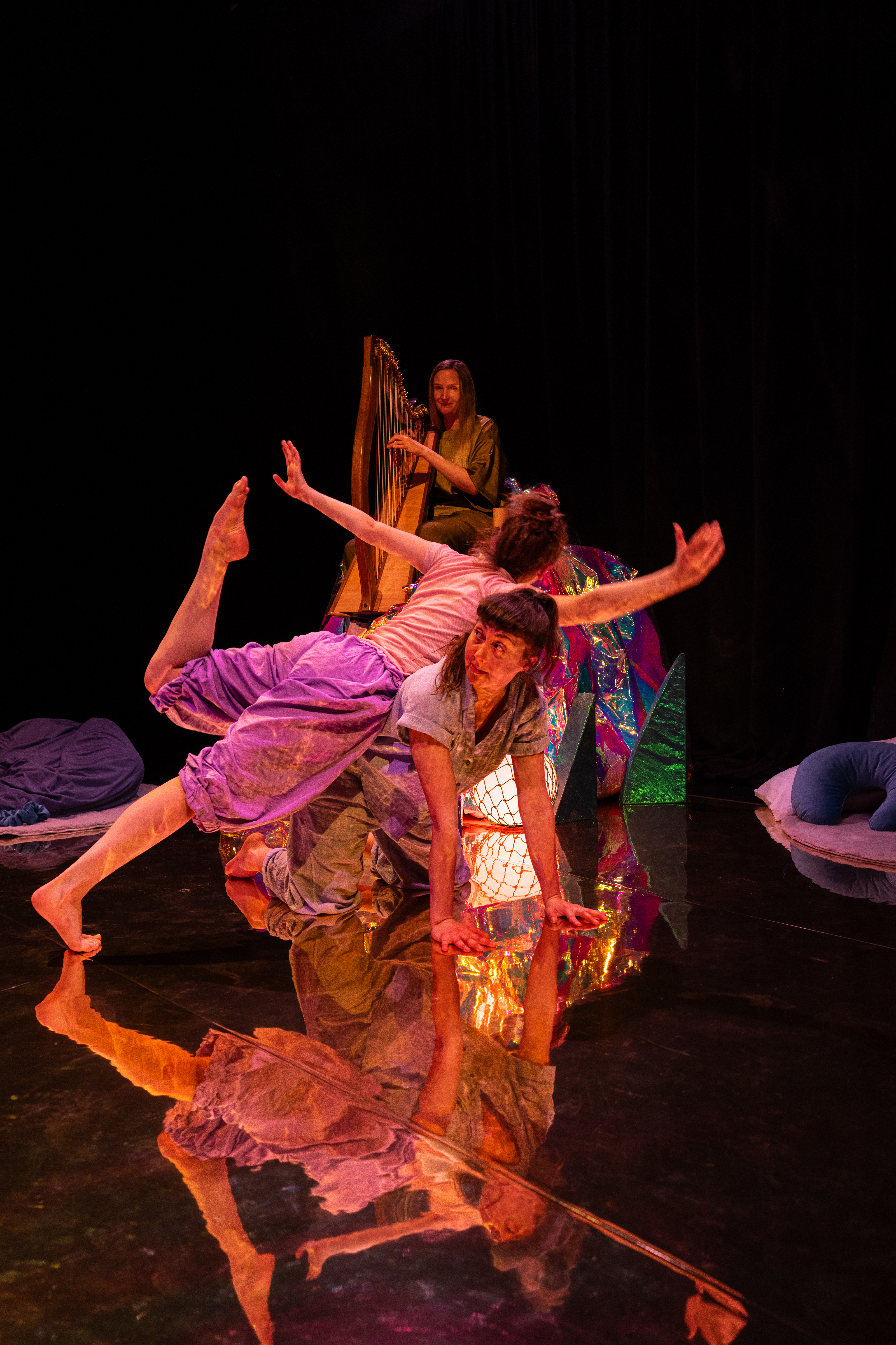 In the foreground two dancers are stacked one on top of the other in a pose. There is someone in the background playing harp.