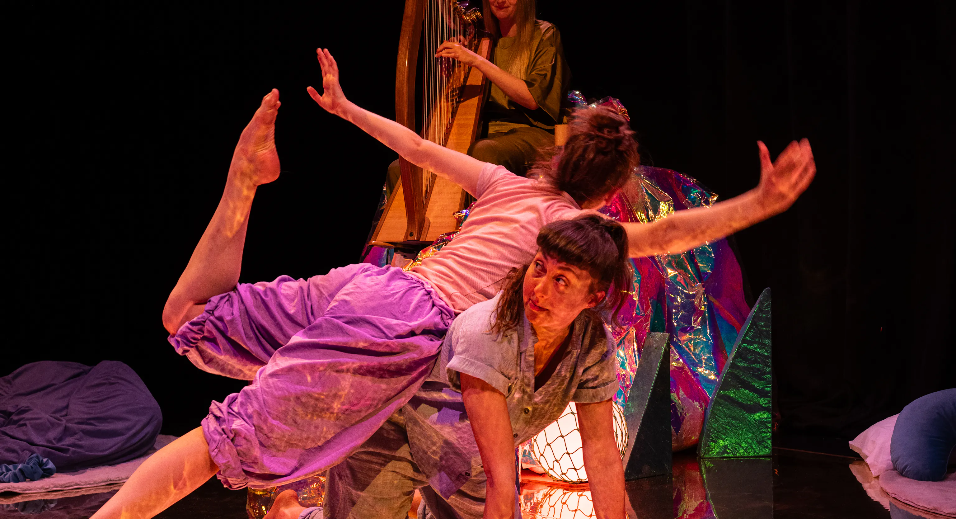 In the foreground two dancers are stacked one on top of the other in a pose. There is someone in the background playing harp.